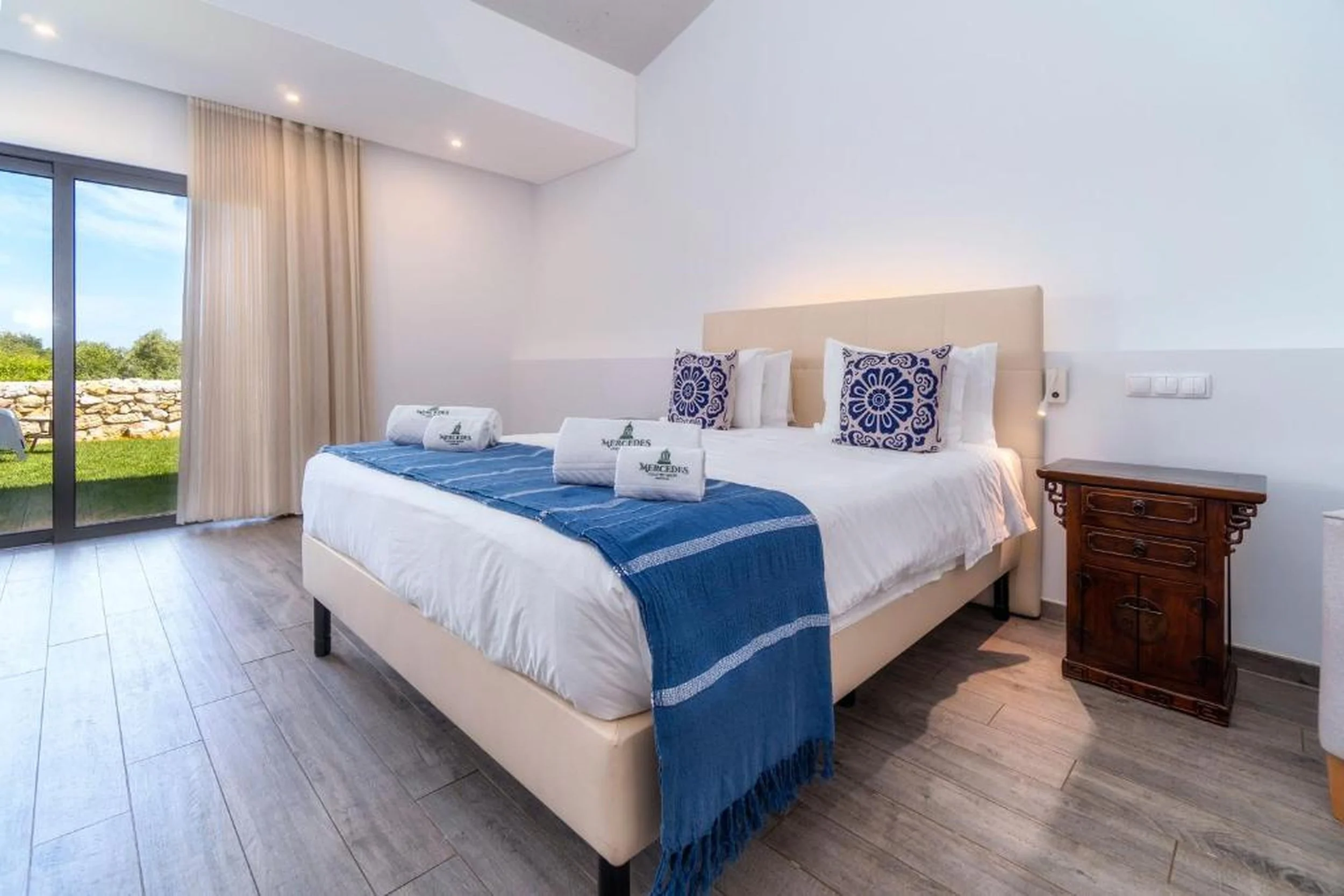 A bright, modern bedroom with a large bed, blue and white bedding, and decorative pillows. There are windows with cream curtains, a wooden nightstand, and view of greenery outside.