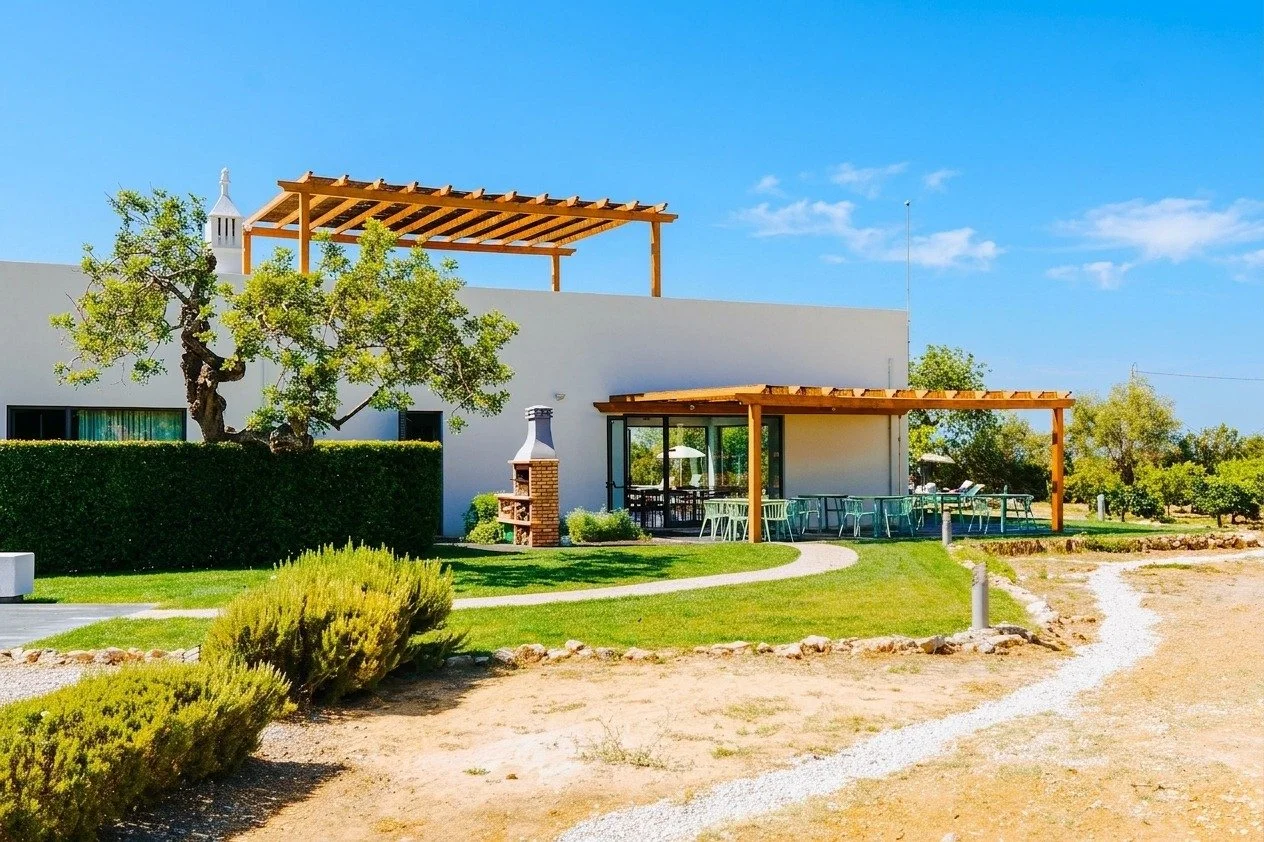 A modern white house with outdoor patio and pergolas, surrounded by green grass, shrubs, and trees, under a clear blue sky.