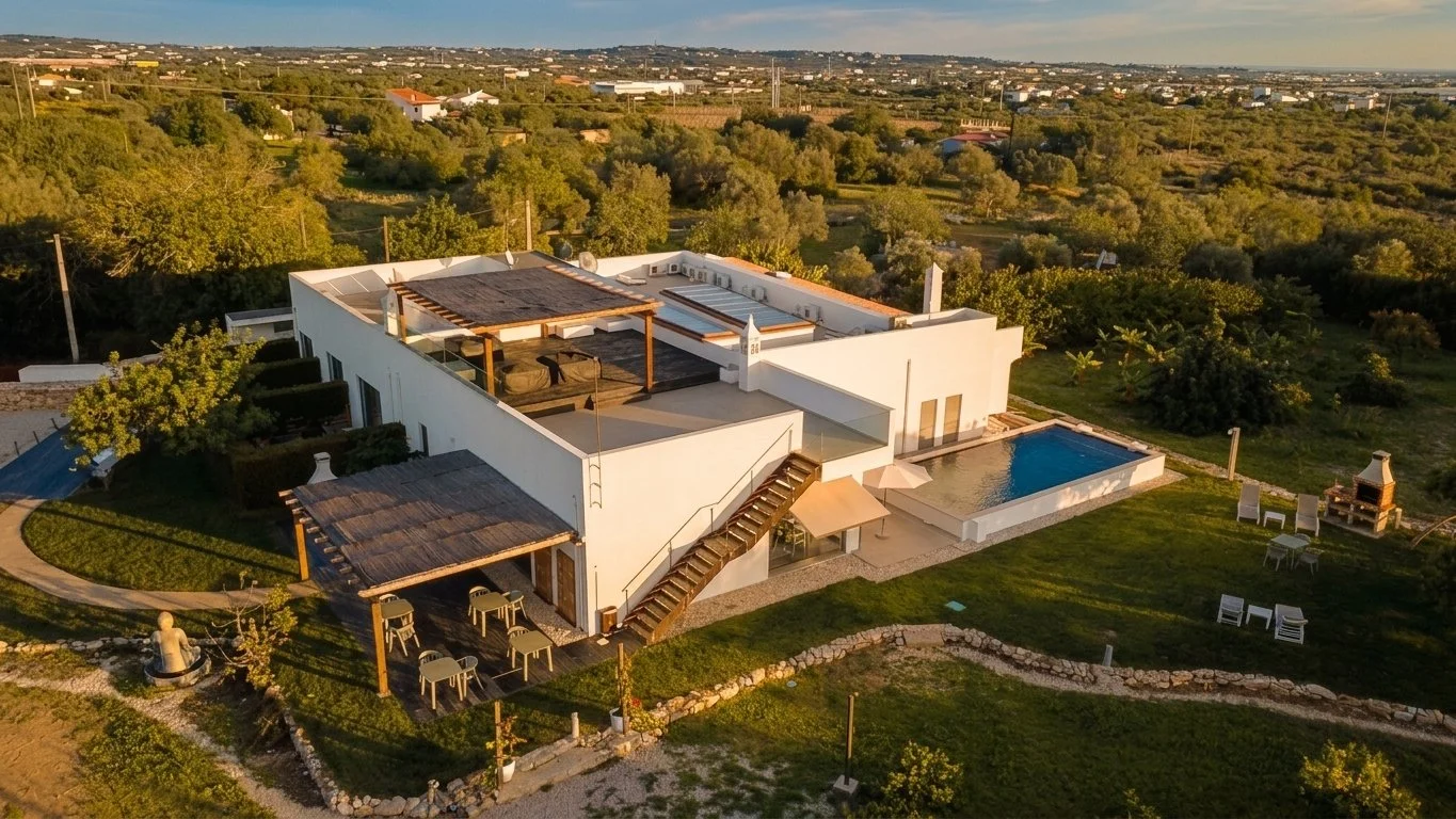 Aerial view of a modern white house with a swimming pool in the backyard, surrounding greenery, outdoor seating, and a landscaped lawn under a clear sky.