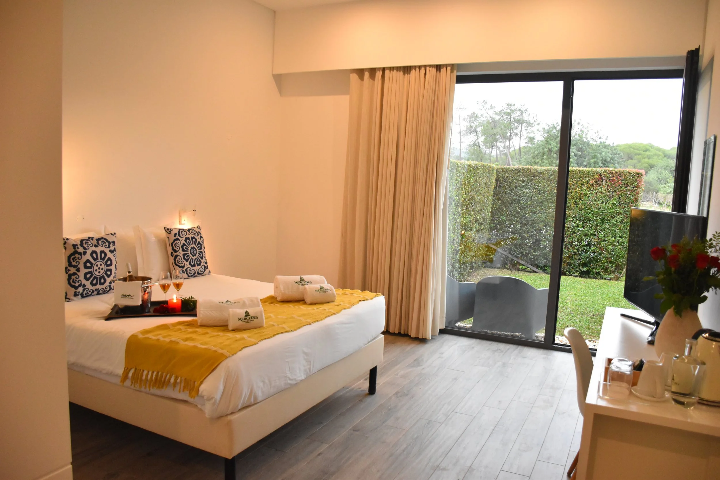 A hotel room with a white bed, black and white decorative pillows, and a gold bed runner. There are towels with the hotel's logo, a tray with beverages, and a lit candle on the bed. Large sliding glass doors open to a lush green outdoor view with trees and a hedge. There is a small table with a flower arrangement and a TV.