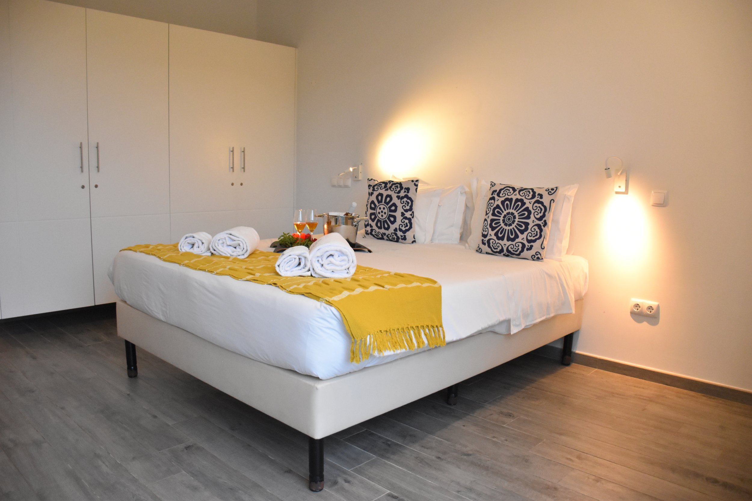 A neatly made hotel bed with white sheets, decorative black and white pillows, a yellow throw blanket at the foot, and rolled white towels on top. There is a tray with wine glasses, a bottle of wine, and a bucket on the bed. The room has a large white wardrobe with silver handles and wall-mounted reading lights.