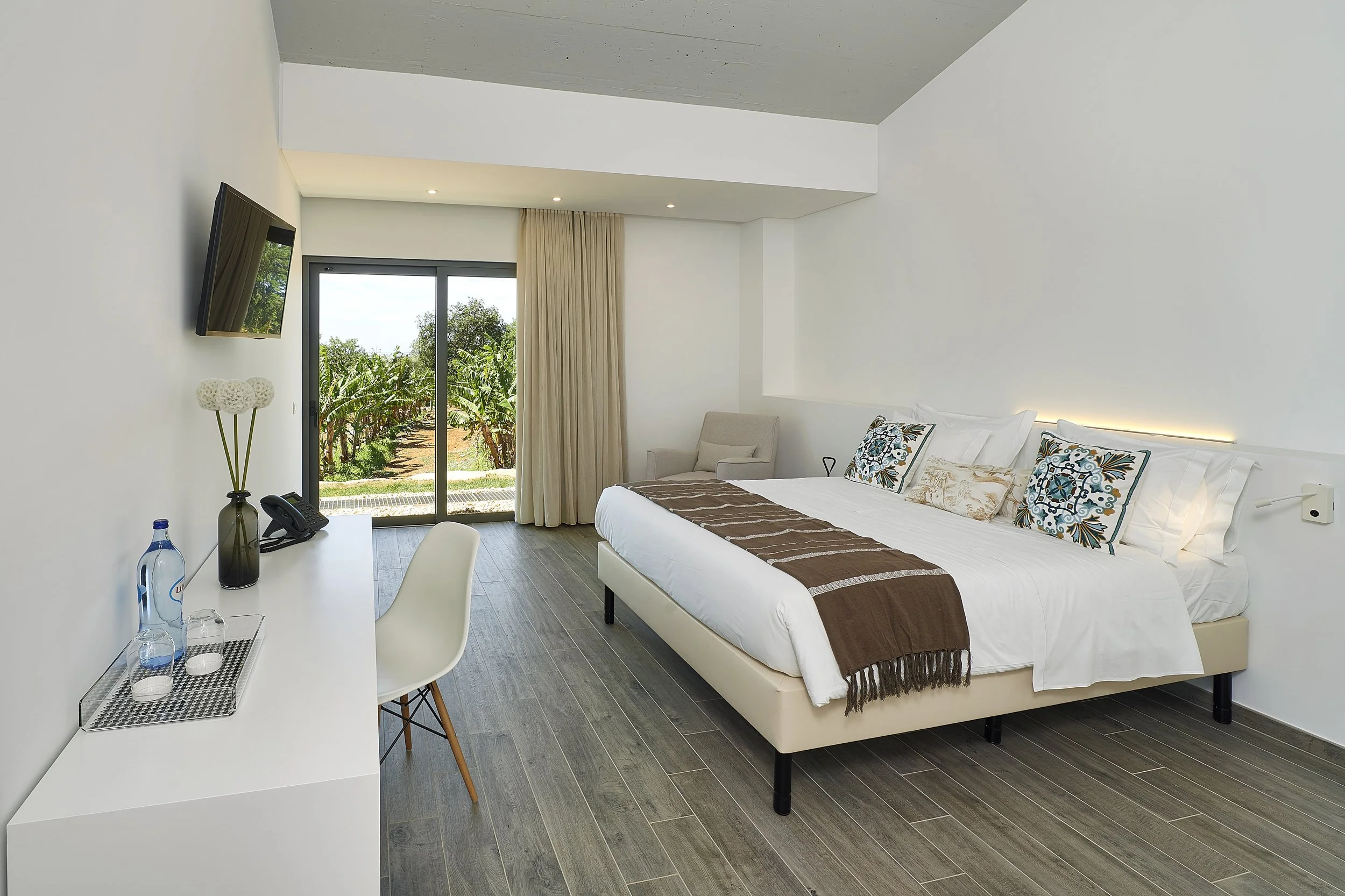 Modern hotel room with a large bed, pillows, and a brown throw blanket, a sitting chair near a window with beige curtains, a wall-mounted TV, and a desk with water bottles, a phone, and decorative vases.