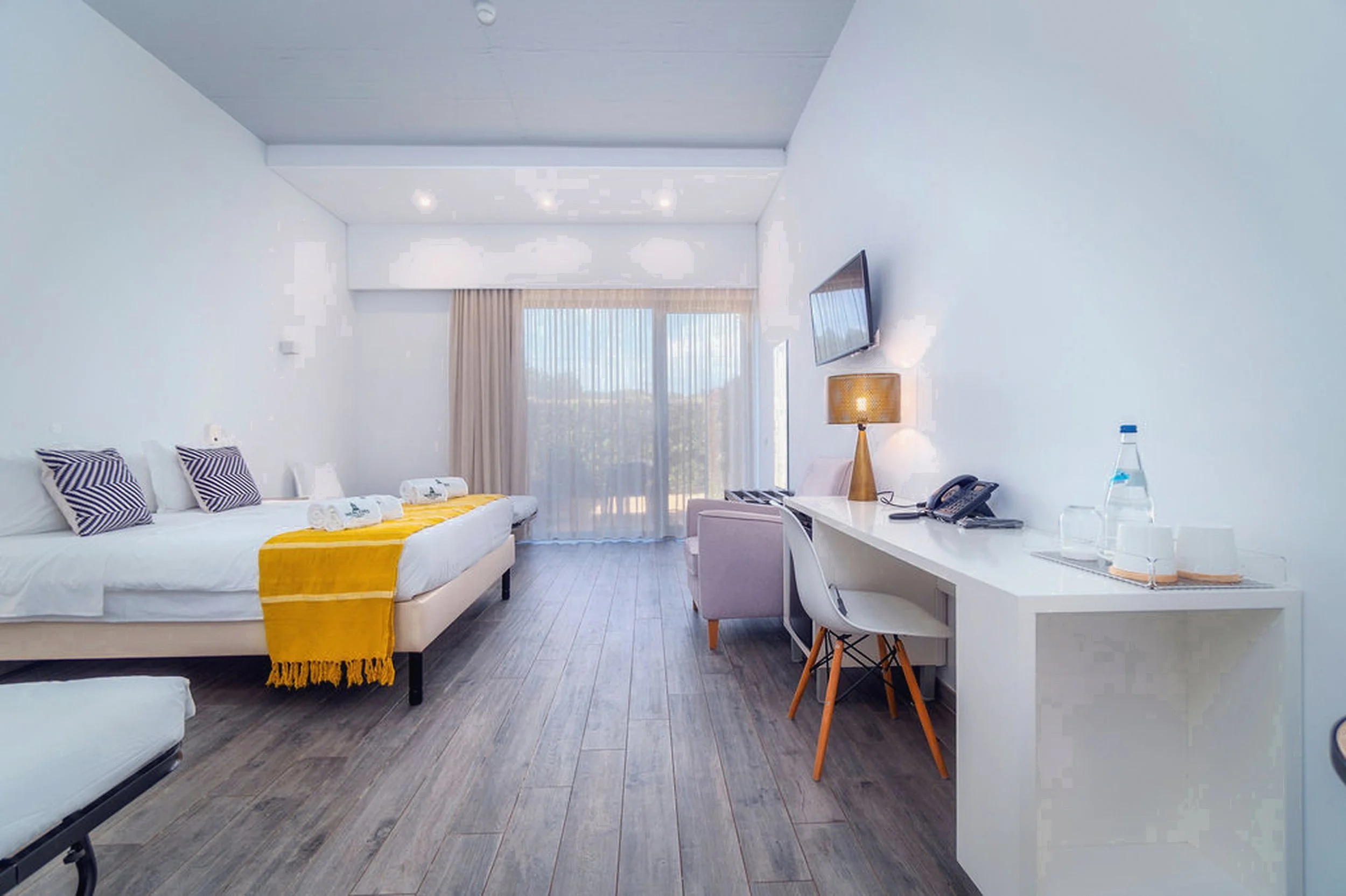 Modern hotel room with two beds, desk, armchair, wall-mounted TV, lamp, and large window with curtains, decorated in white and yellow accents.
