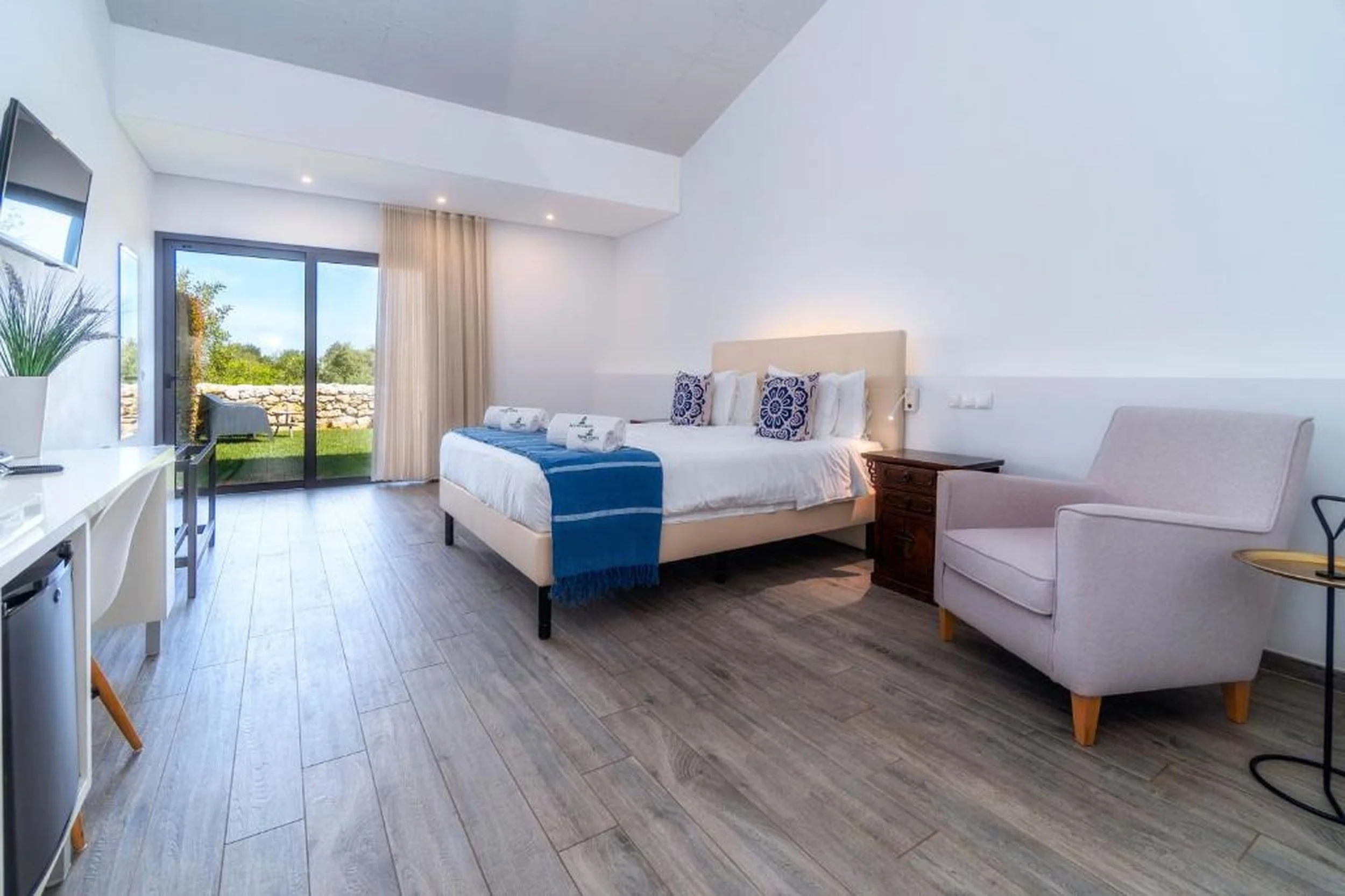 A modern hotel room with a large bed, two rolled towels, and decorative pillows, with access to an outdoor area with grass and stone wall visible through sliding glass doors.