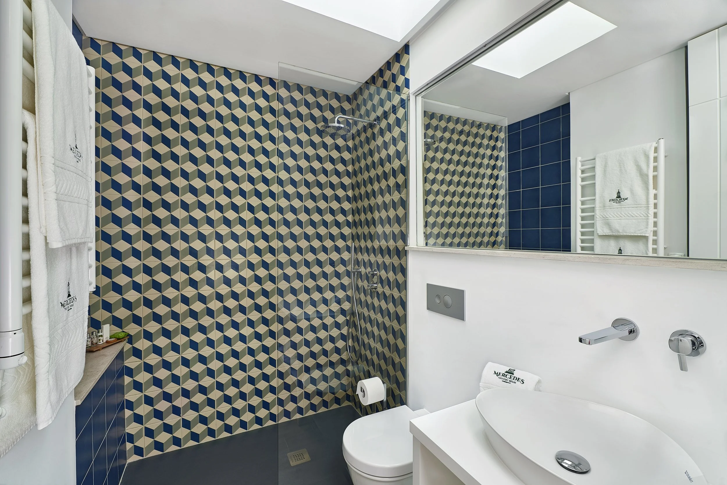 Modern bathroom with geometric patterned wall tiles, white fixtures, shower area, large mirror, towel rack with towels, and skylight.