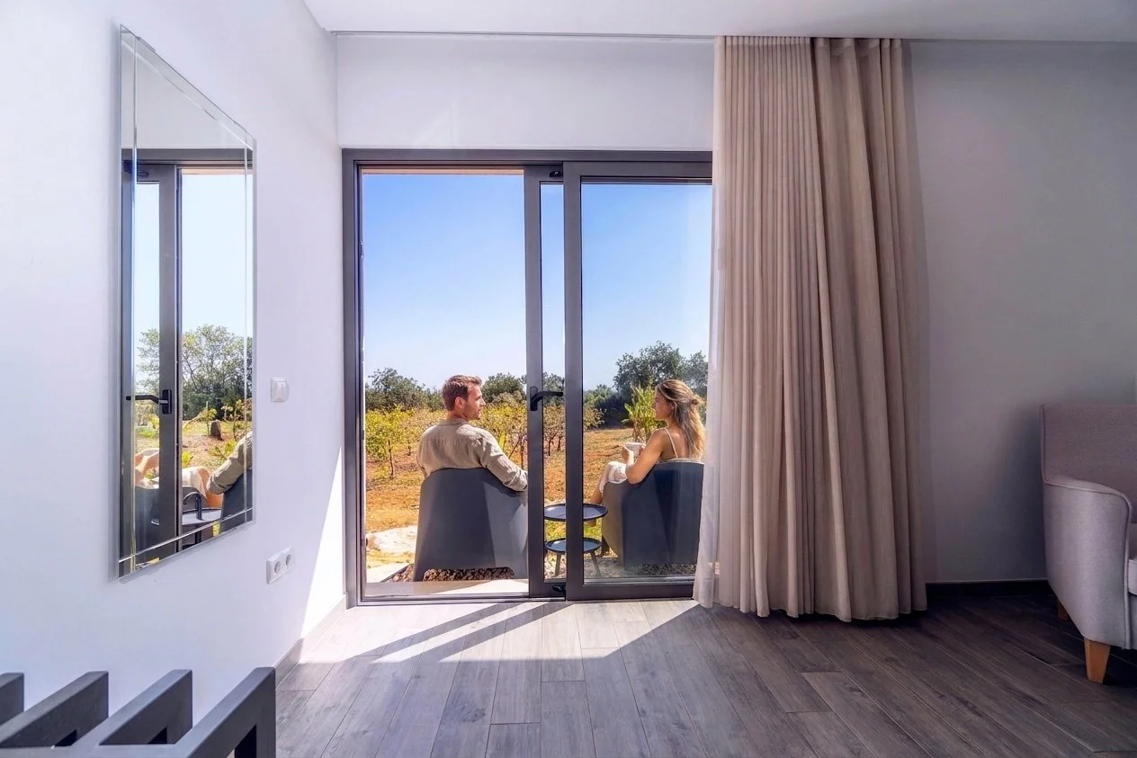 A view from inside a modern room looking out through glass sliding doors to two people sitting outside on chairs, enjoying an outdoor scene of trees and grass on a sunny day.