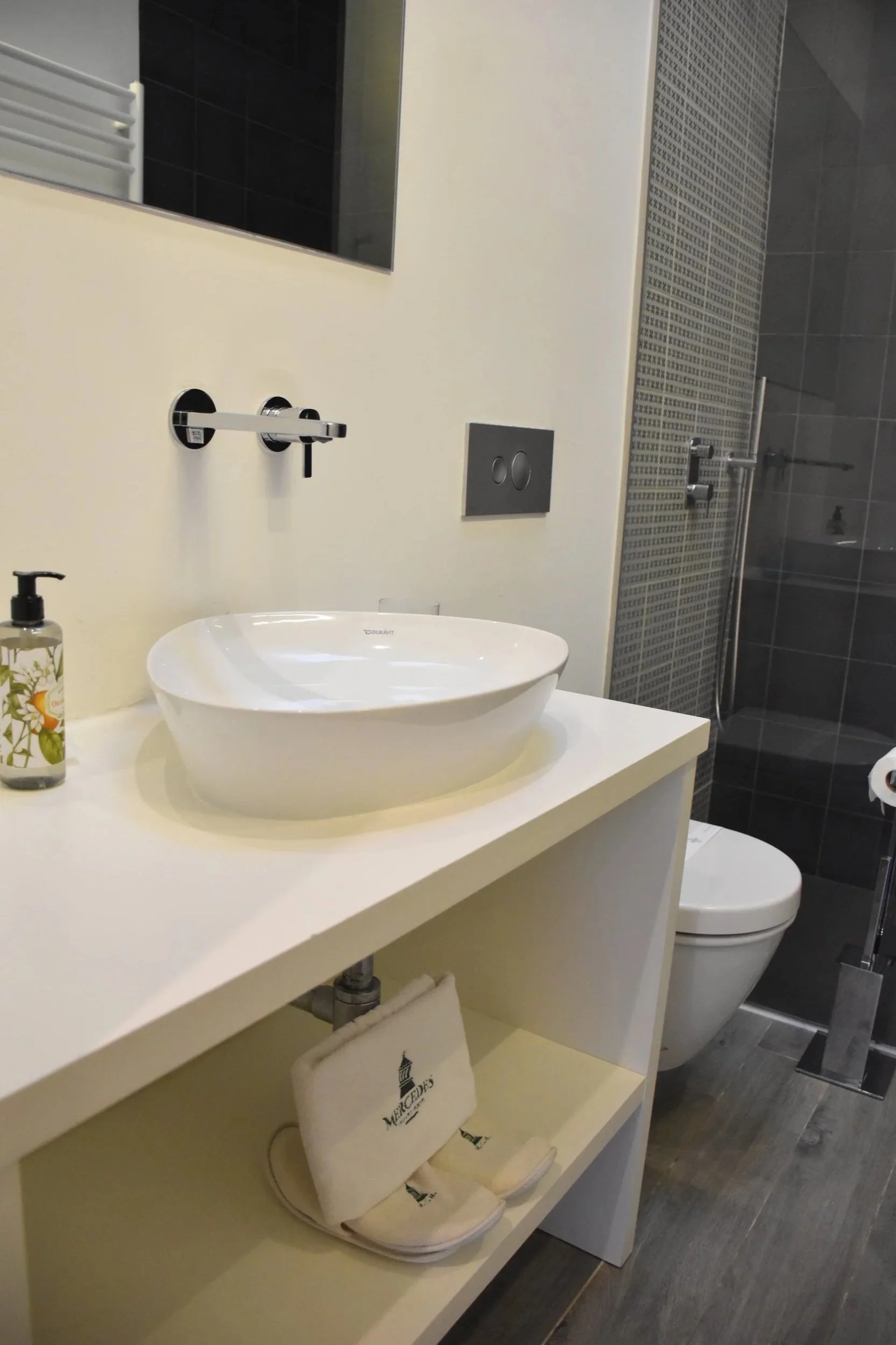 Modern bathroom with white sink on a white vanity, toilet, large mirror, soap dispenser, and a walk-in shower with glass door and dark tiles.