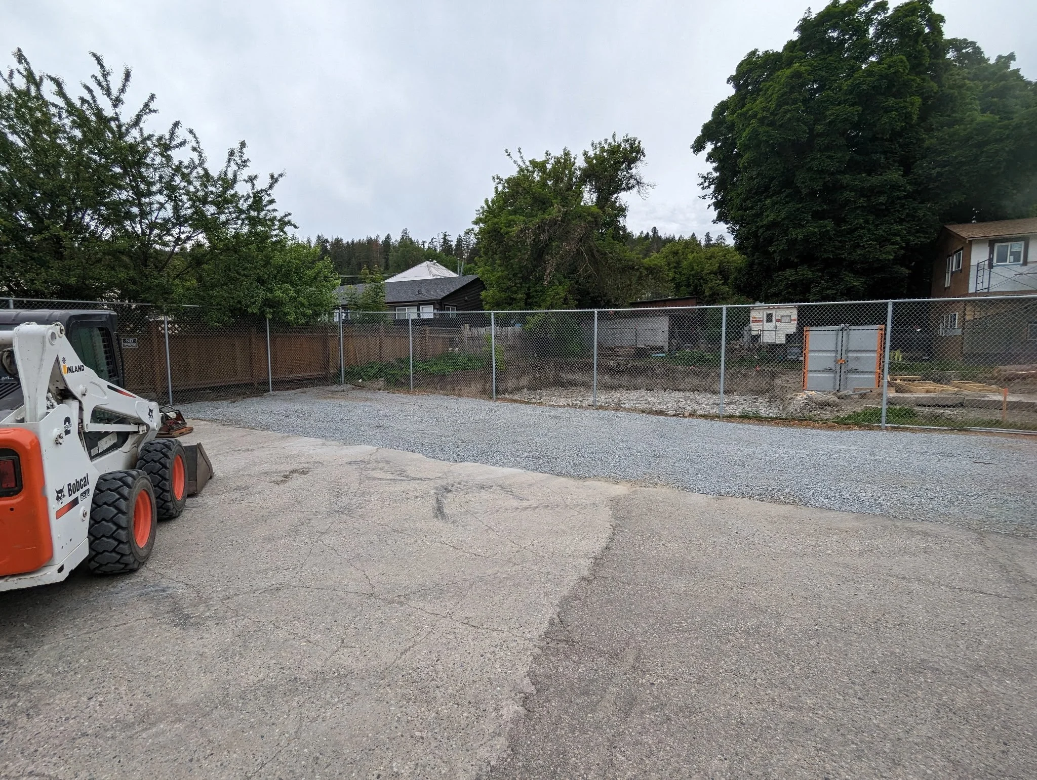 Construction site with gravel and dirt, surrounded by a chain-link fence, with a Bobcat skid-steer loader parked on cracked asphalt in the foreground and residential houses with trees in the background.