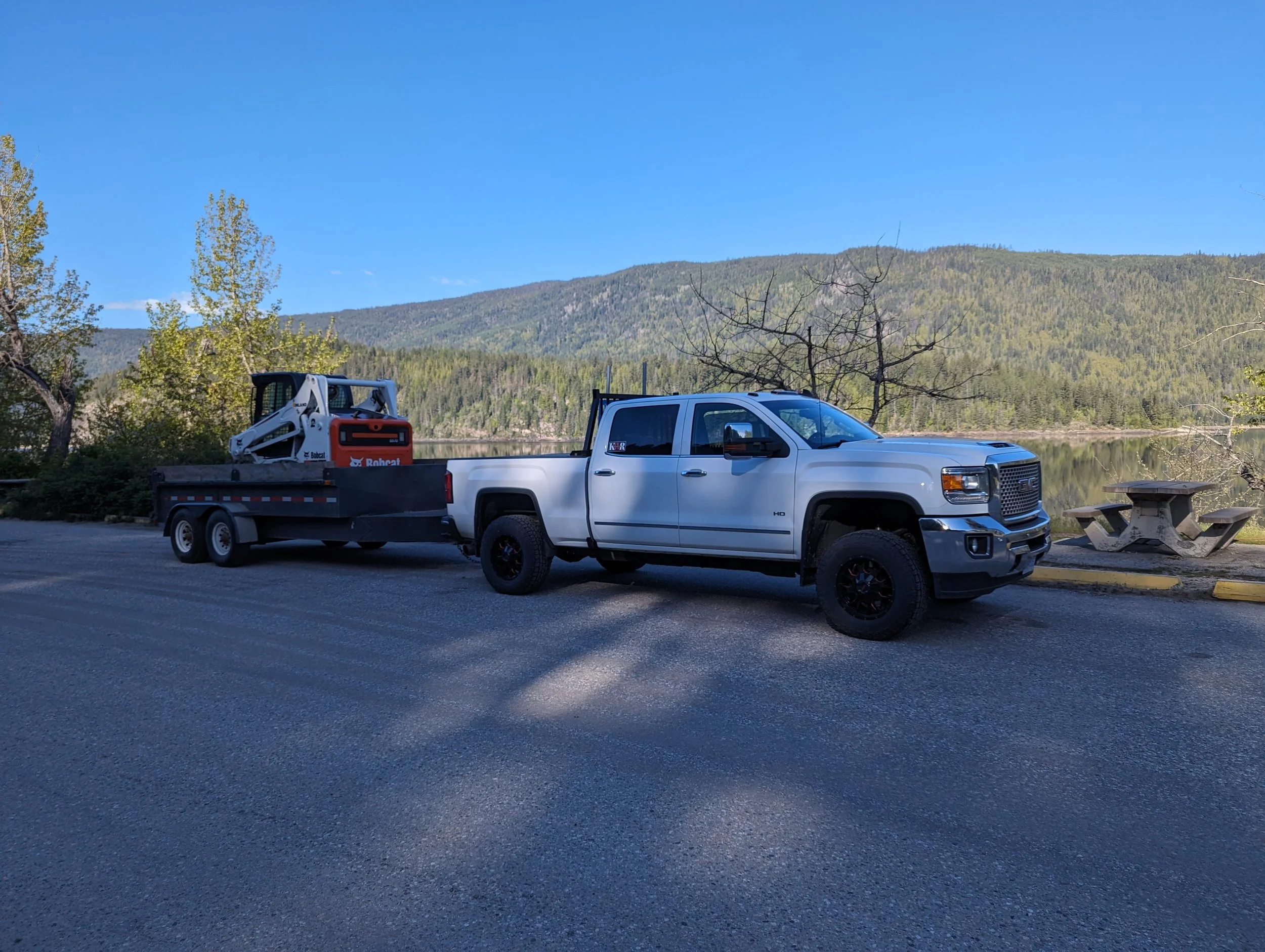 A white pickup truck with a flatbed trailer attached, carrying a Bobcat compact loader, parked near a lake with trees and hills in the background under a clear blue sky.