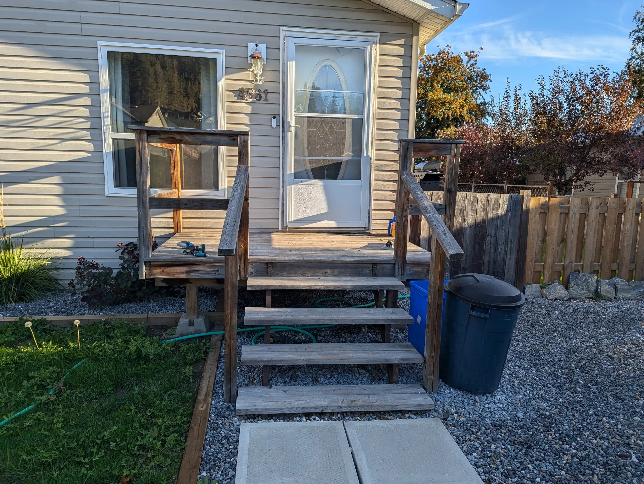 Wooden stairs leading to a back door of a house with a small porch. The house has beige siding, a window, and a white door with an oval window. There is a trash bin, a blue recycling bin, and some gardening tools on the porch. Surrounding the house are trees with autumn foliage and a gravel pathway.