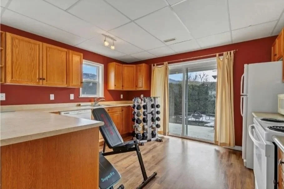 Kitchen with wooden cabinets, a window, a sliding glass door with curtains leading outside, a white refrigerator, a white stove, a black chair, and a folded drying rack, with a view of an outdoor patio.