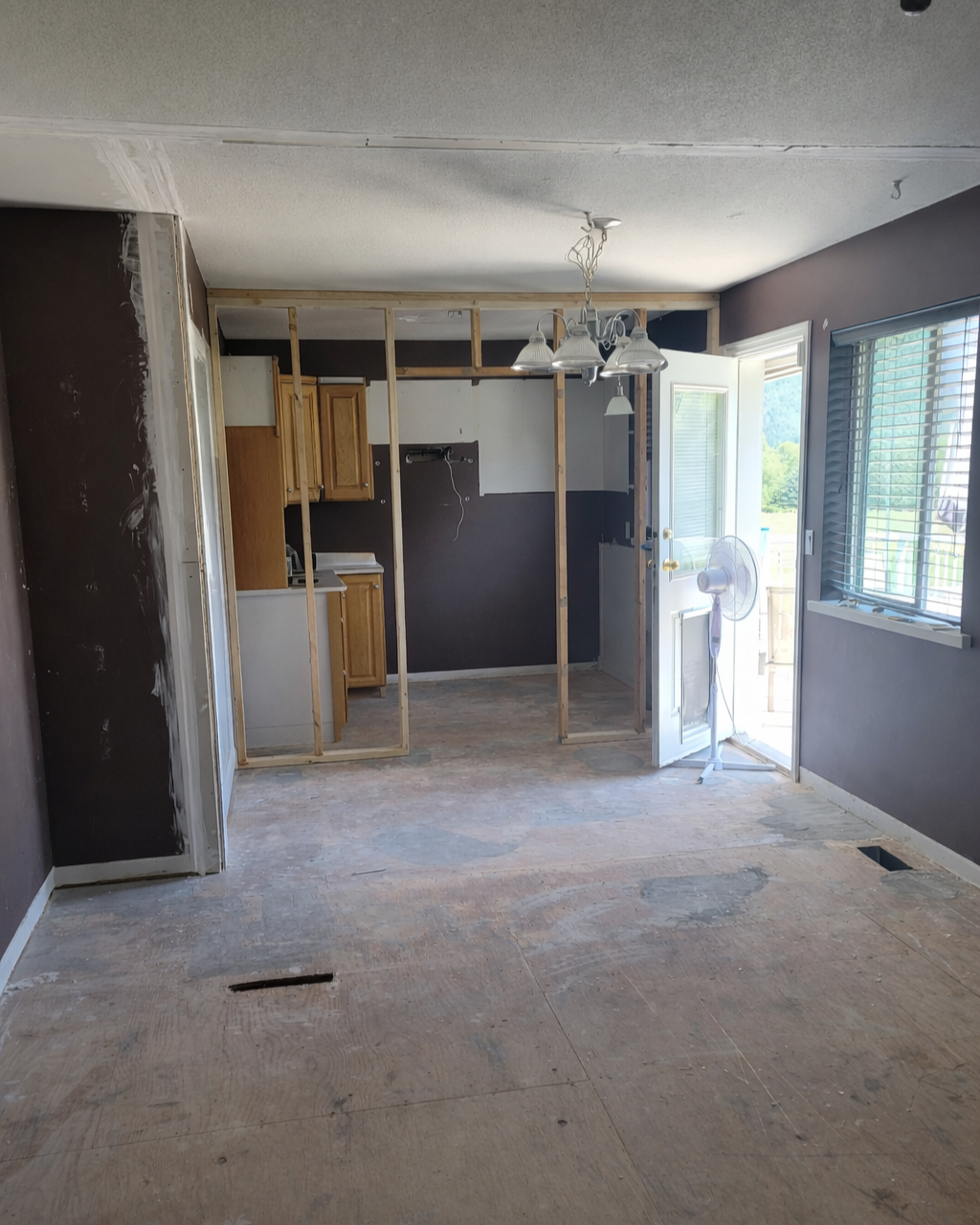 Room under renovation with exposed wooden framing, partially installed kitchen cabinets, a fan near the door, and a chandelier hanging from the ceiling.