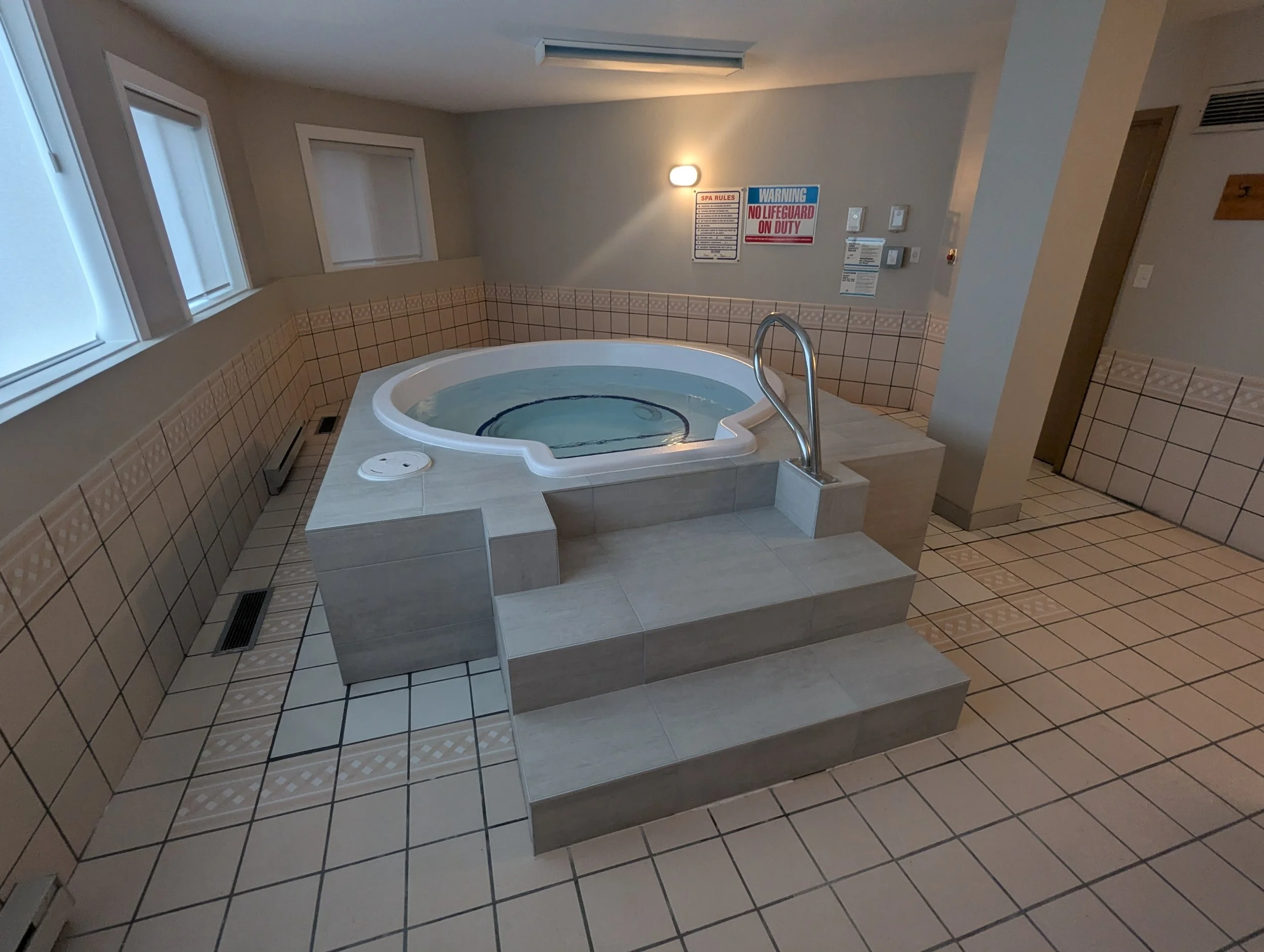 Indoor hot tub with water, surrounded by beige tiles, three steps leading up to it, and a metallic handrail.