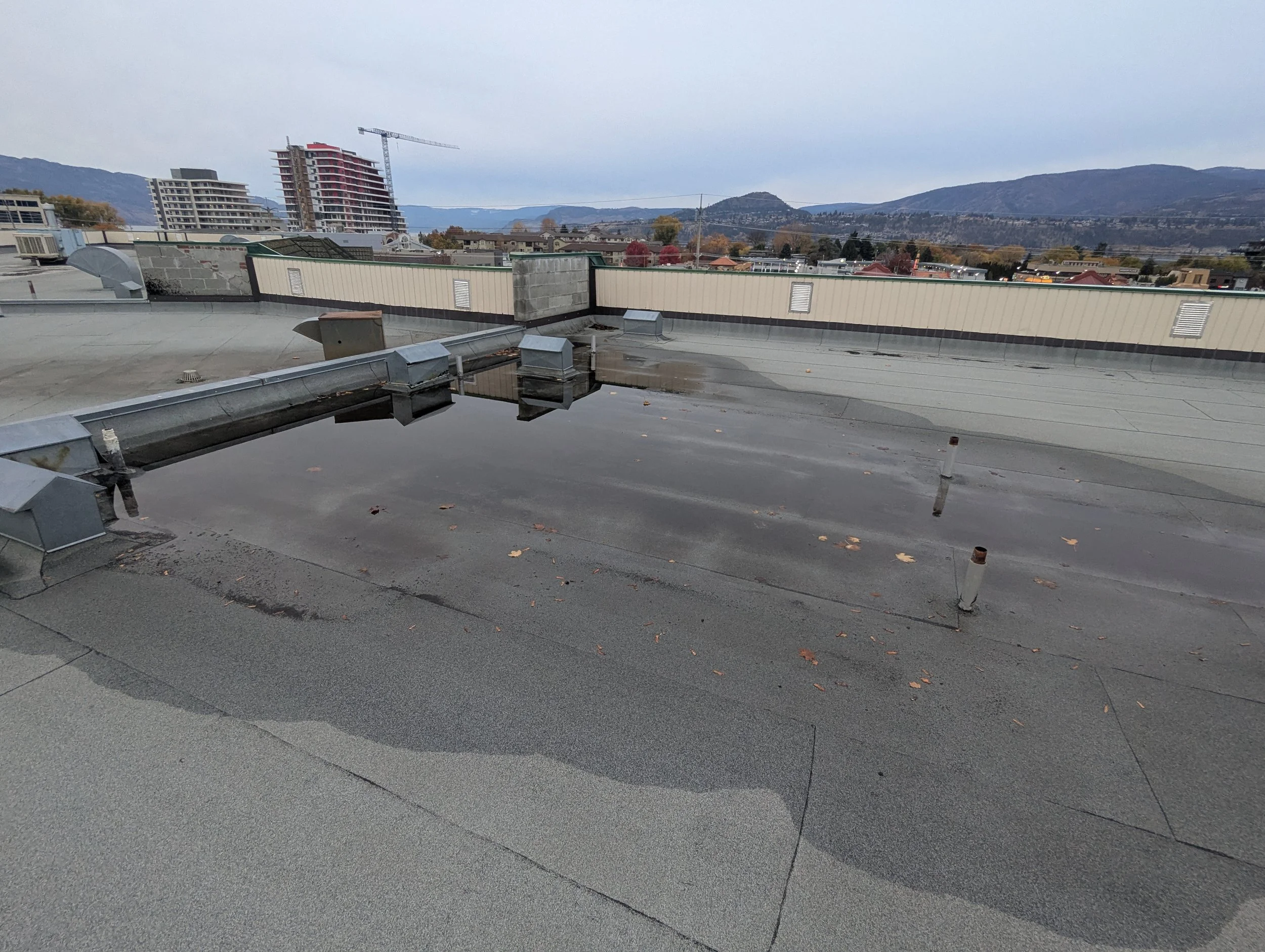 View of a cityscape from a rooftop with roofing materials, vents, and a water puddle, with mountains in the background under an overcast sky.