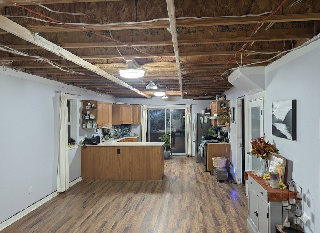 Living space with unfinished ceiling exposing wooden beams and insulation, hardwood floors, and a kitchen area with wooden cabinets and a countertop. There are windows with curtains, a sliding glass door at the back, and various household items and plants.