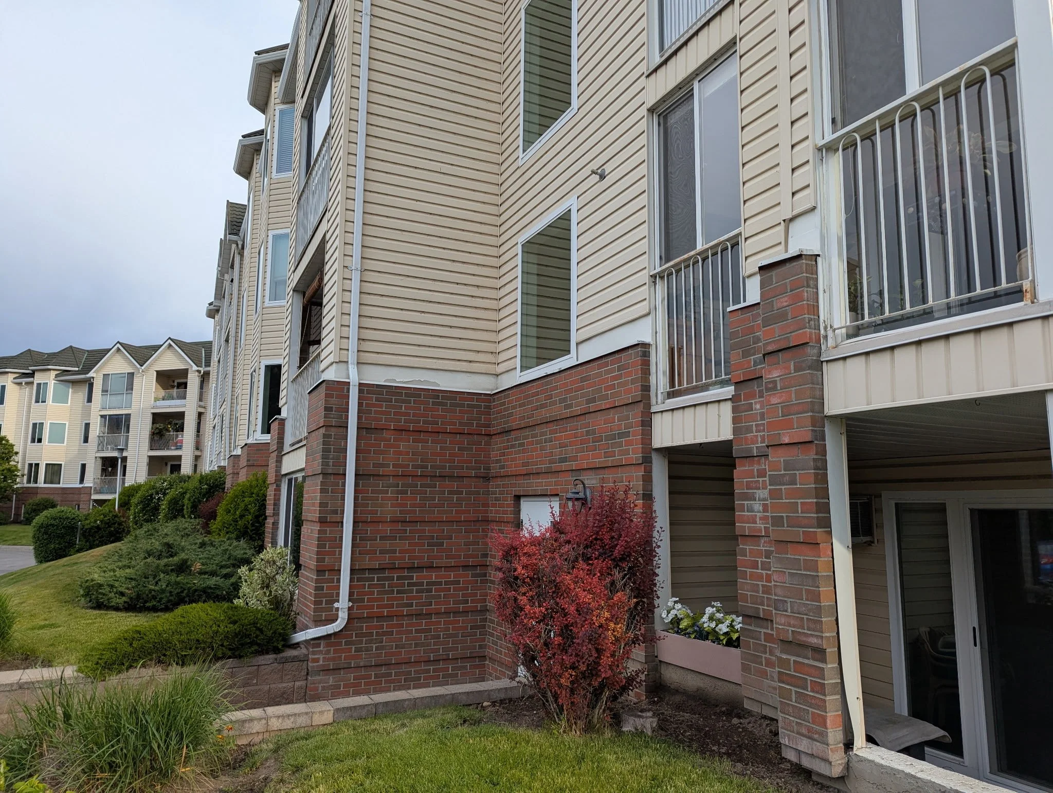 View of a multi-story apartment building with beige siding, brick accents, balconies with white railings, and a landscaped lawn with shrubs and a red bush, under a cloudy sky.