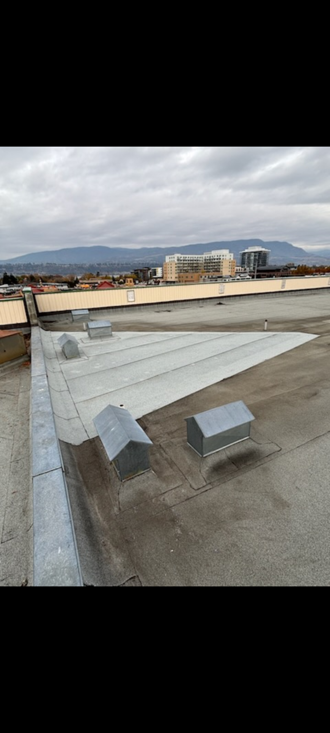 View of a rooftop with multiple vent pipes and a low wall, overlooking a cityscape with buildings and mountains under a cloudy sky.