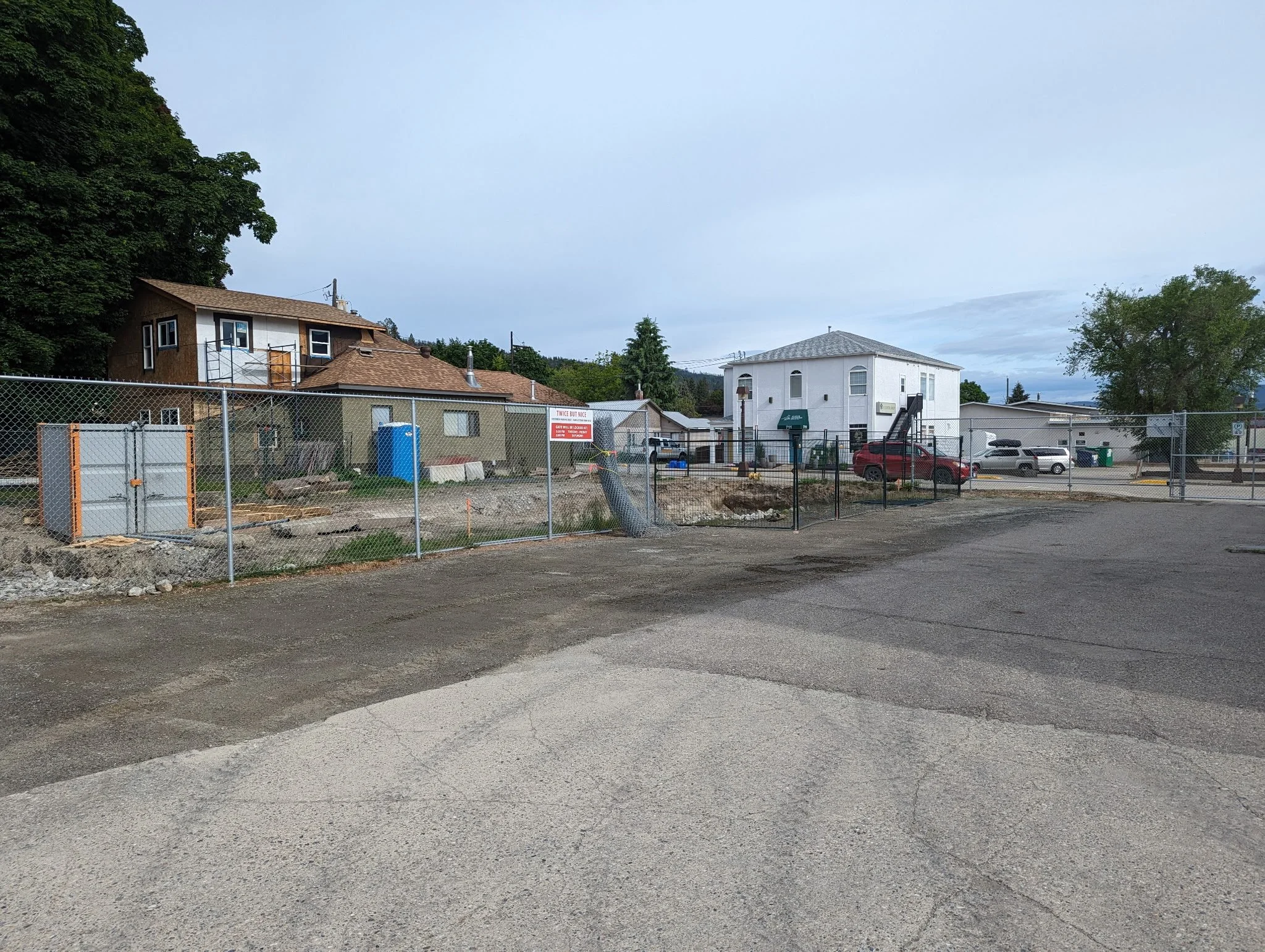 A construction site enclosed by a chain-link fence with a sign, dirt and construction materials in front of residential houses, parked cars, and trees in the background.