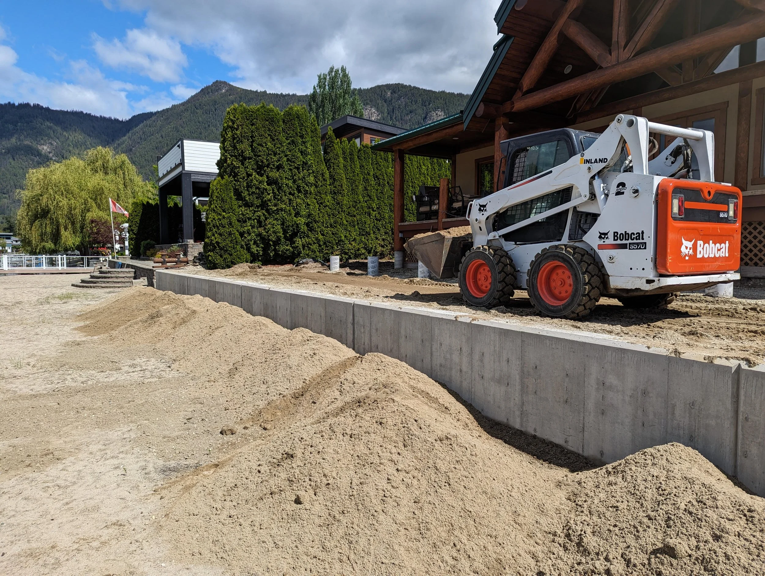 Construction site with piles of sand, a Bobcat skid-steer loader, a concrete wall, and a house with a wooden porch surrounded by green trees and mountains in the background.