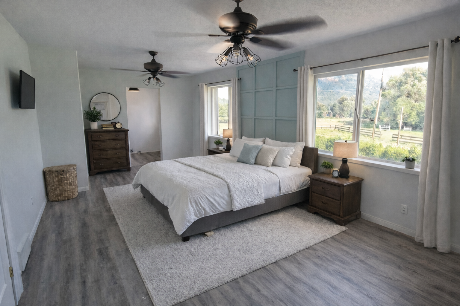 A bedroom with a large bed, white bedding, and multiple pillows, flanked by two wooden nightstands with lamps. Large windows offer a view of greenery and mountains outside. The room has white walls, a light wood floor, and ceiling fans.
