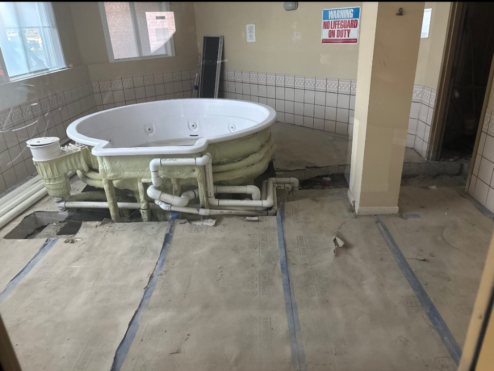 A hot tub under renovation in a room with beige walls, tiled walls, and a window. Construction materials and pipes are visible on the floor.