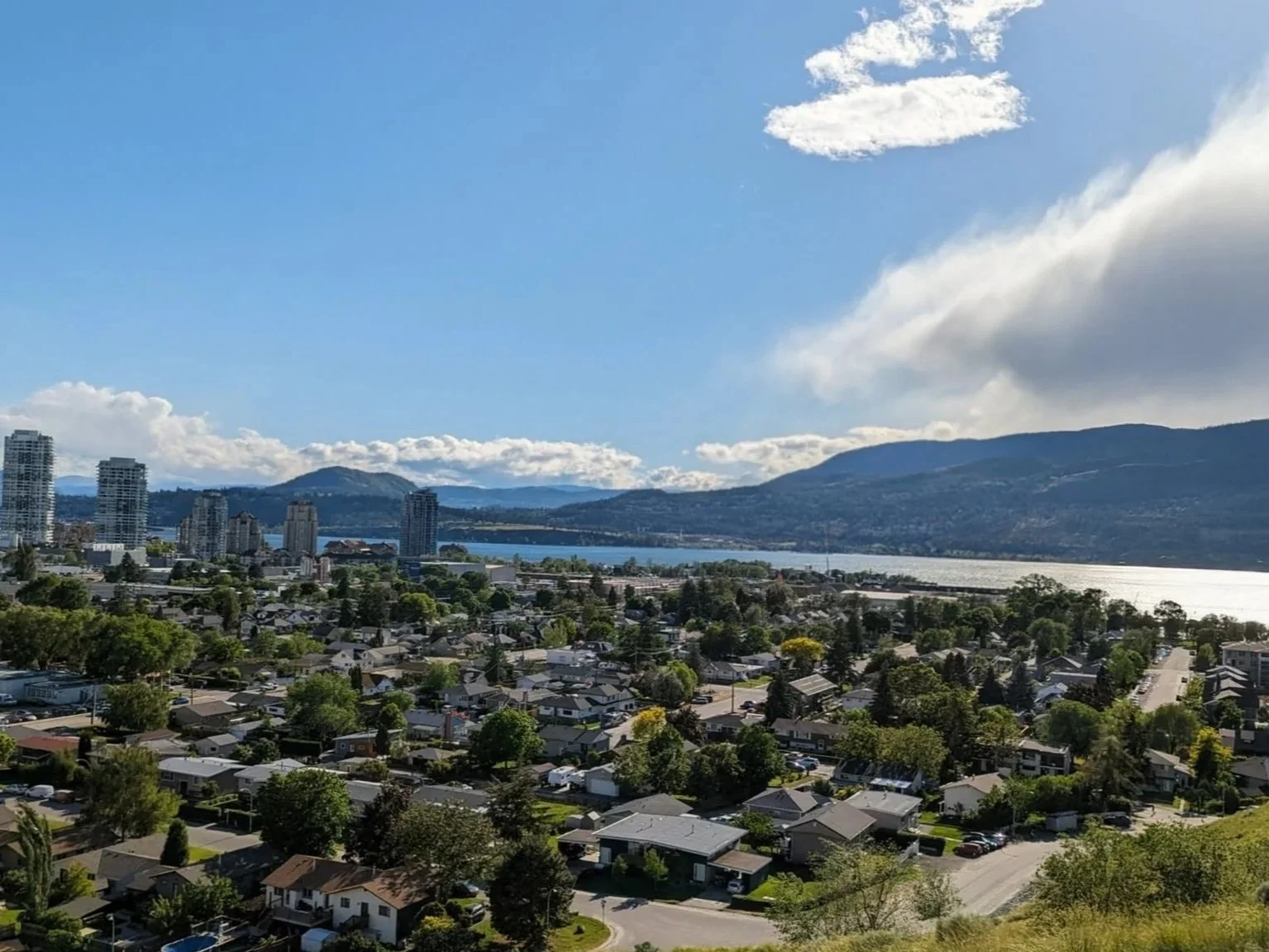 A cityscape with residential houses and a few high-rise buildings, set against a backdrop of mountains and a body of water, under a partly cloudy sky.
