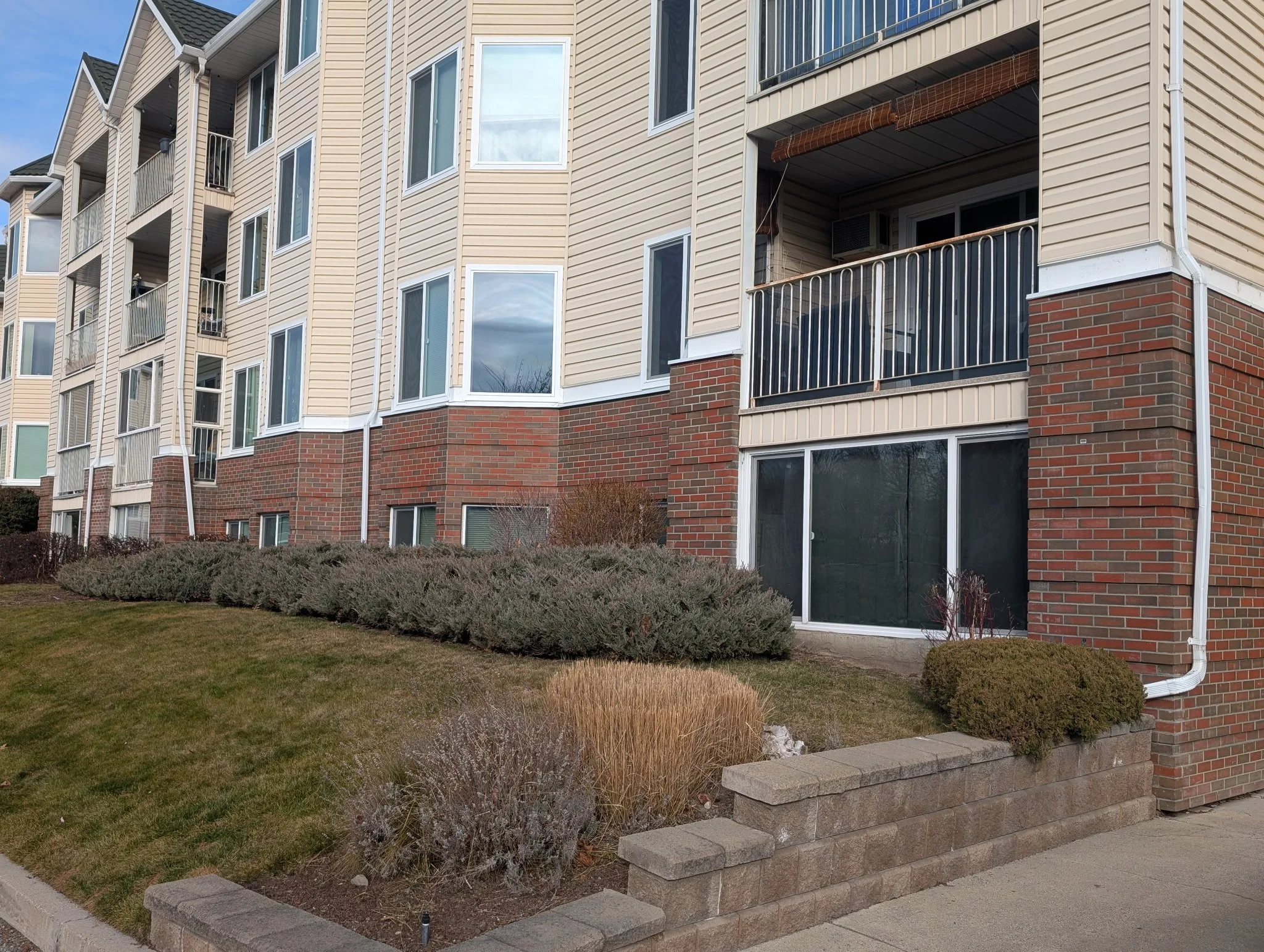 A multi-story apartment building with beige siding and brick accents, featuring balconies and large windows. The building is next to a landscaped area with shrubs, bushes, and a sloped grass lawn, bordered by a small brick wall and sidewalk.