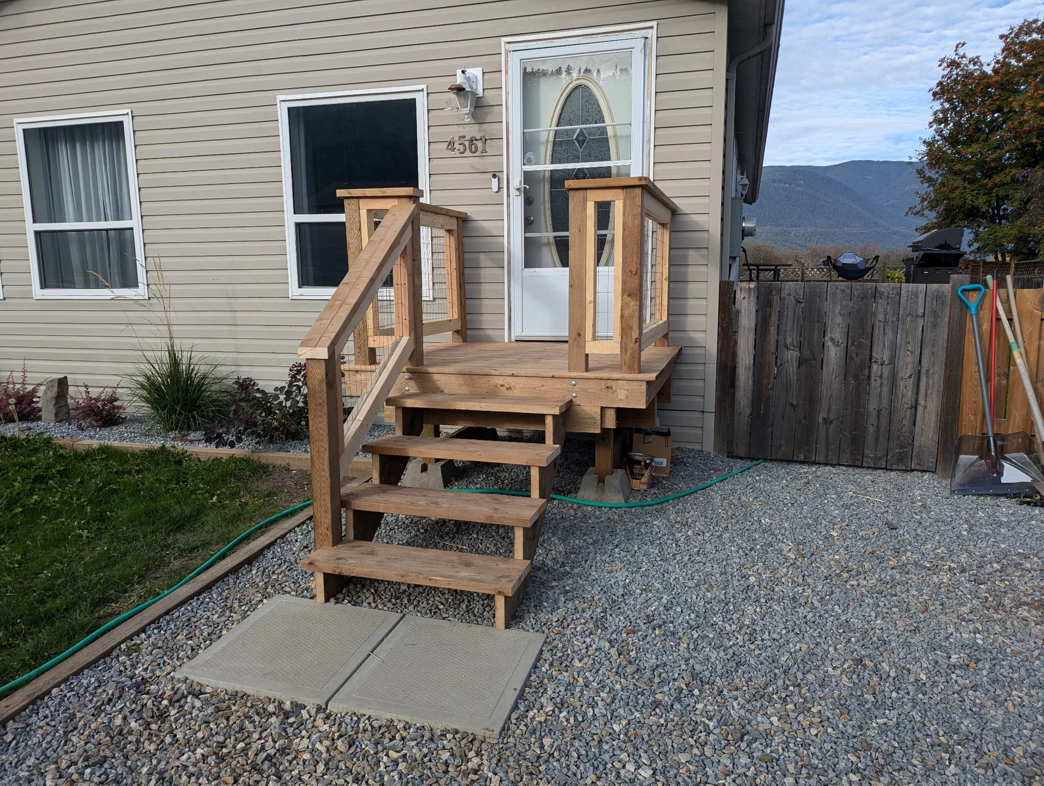 New wooden deck with stairs and railings attached to the back of a beige house with two windows and a glass door with decorative oval window. Gravel yard with tools and a garden bed with plants and shrubs.