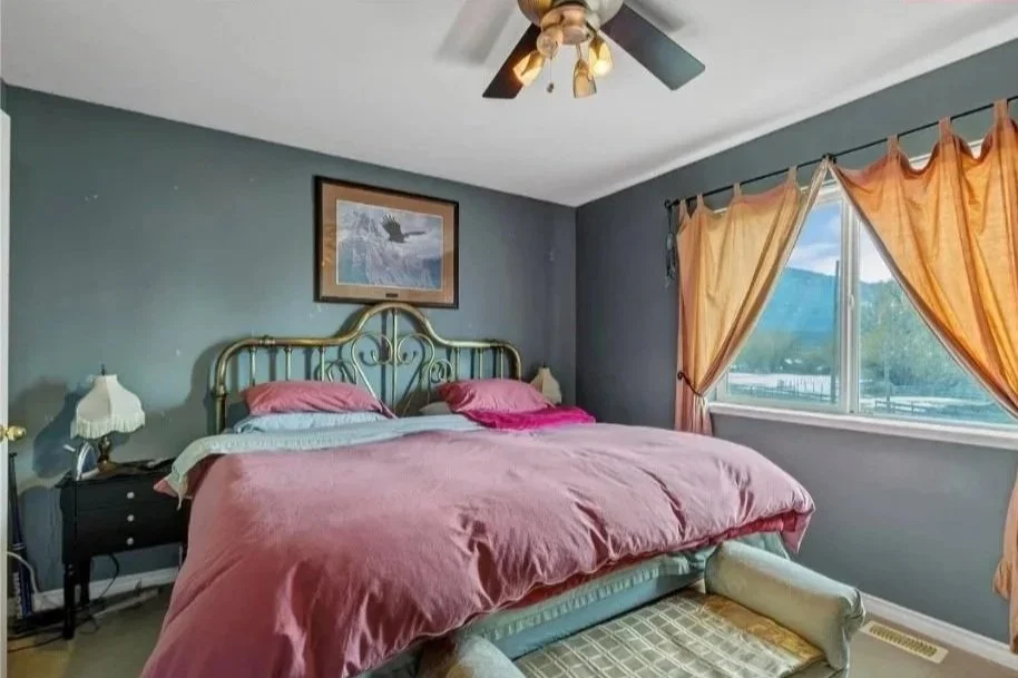 A bedroom with a bed, pink bedding, a nightstand, lamp, window with orange curtains, ceiling fan, and framed wall art of a whale.