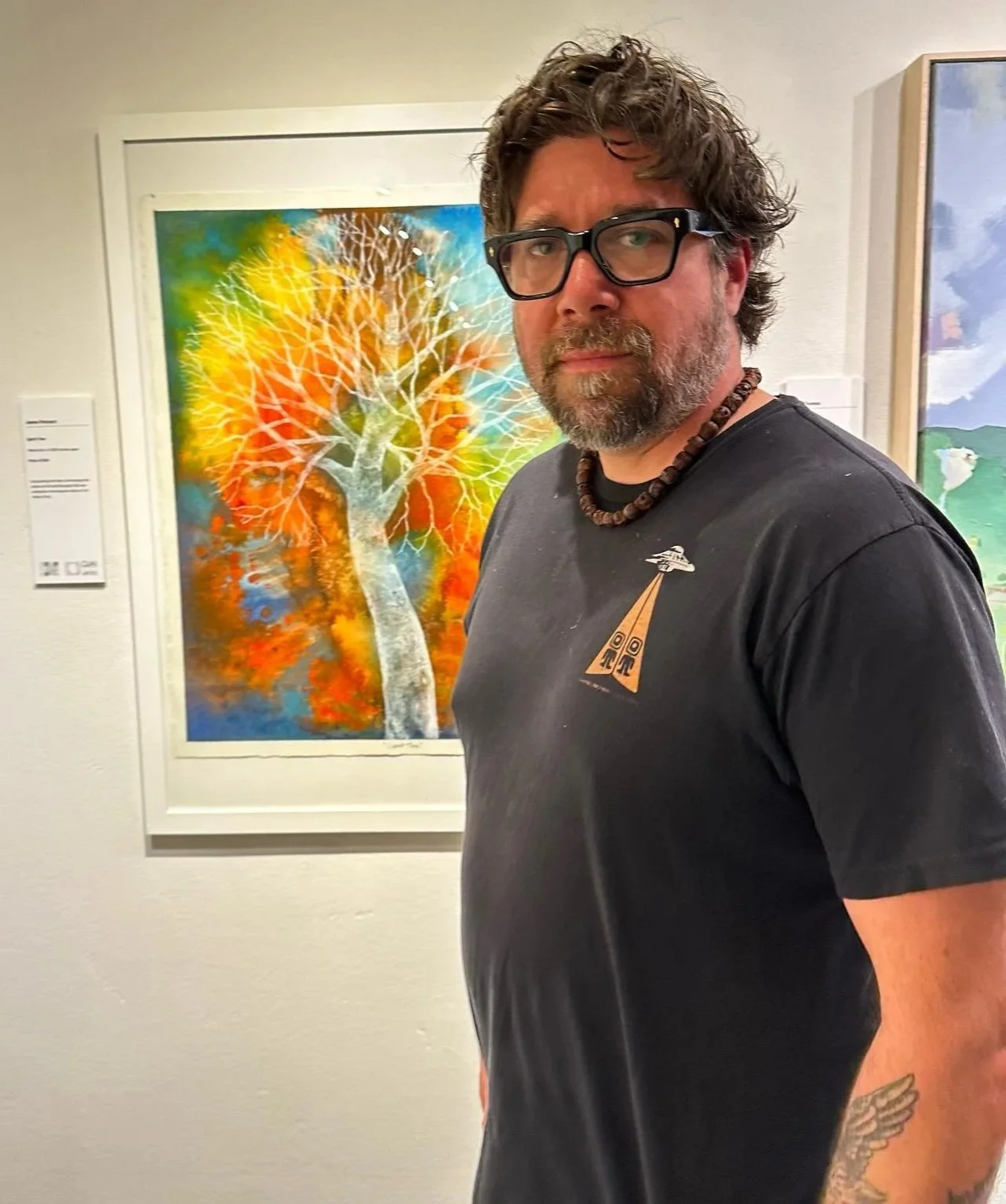 Man with glasses and a beard wearing a black t-shirt with a colorful design, standing in front of vibrant abstract art of a white-outlined tree against a watercolor background in an art gallery.