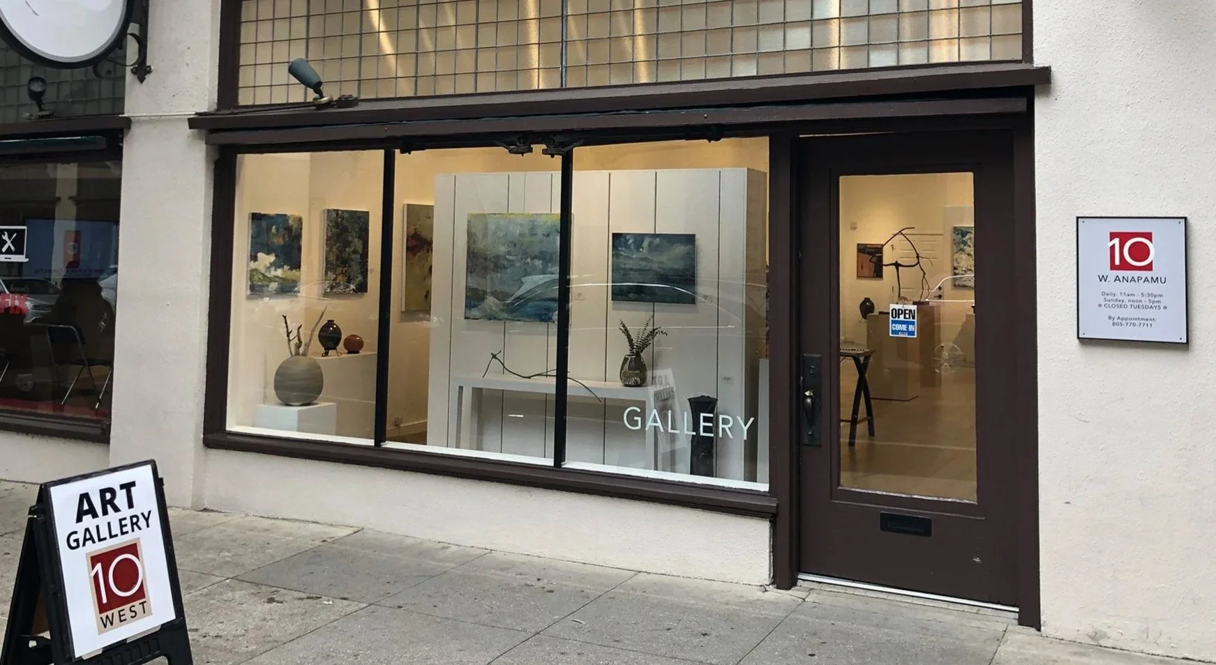 Art gallery storefront with large window displaying paintings and sculptures, entrance door with "Open" sign, sidewalk sign advertising art gallery, and street sidewalk.