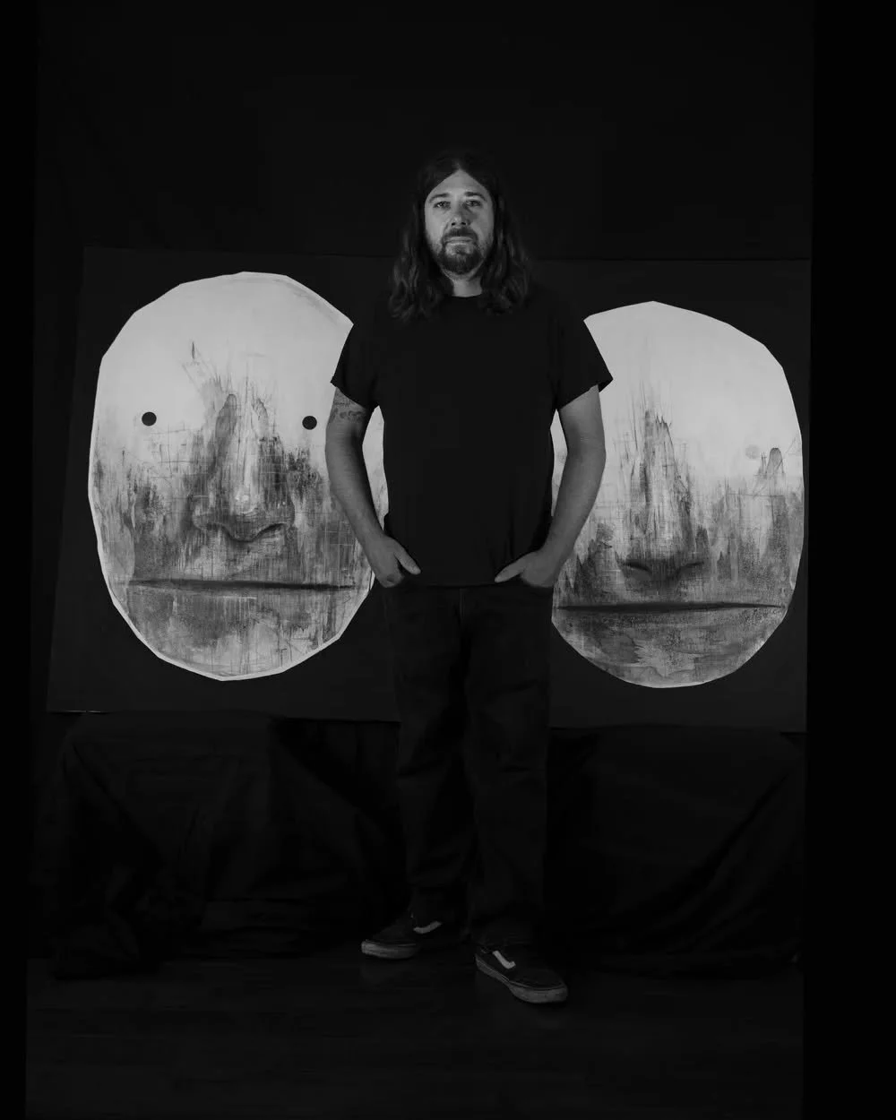 A man with long hair and a beard standing in front of a large, abstract painting with two faces. The image is in black and white.