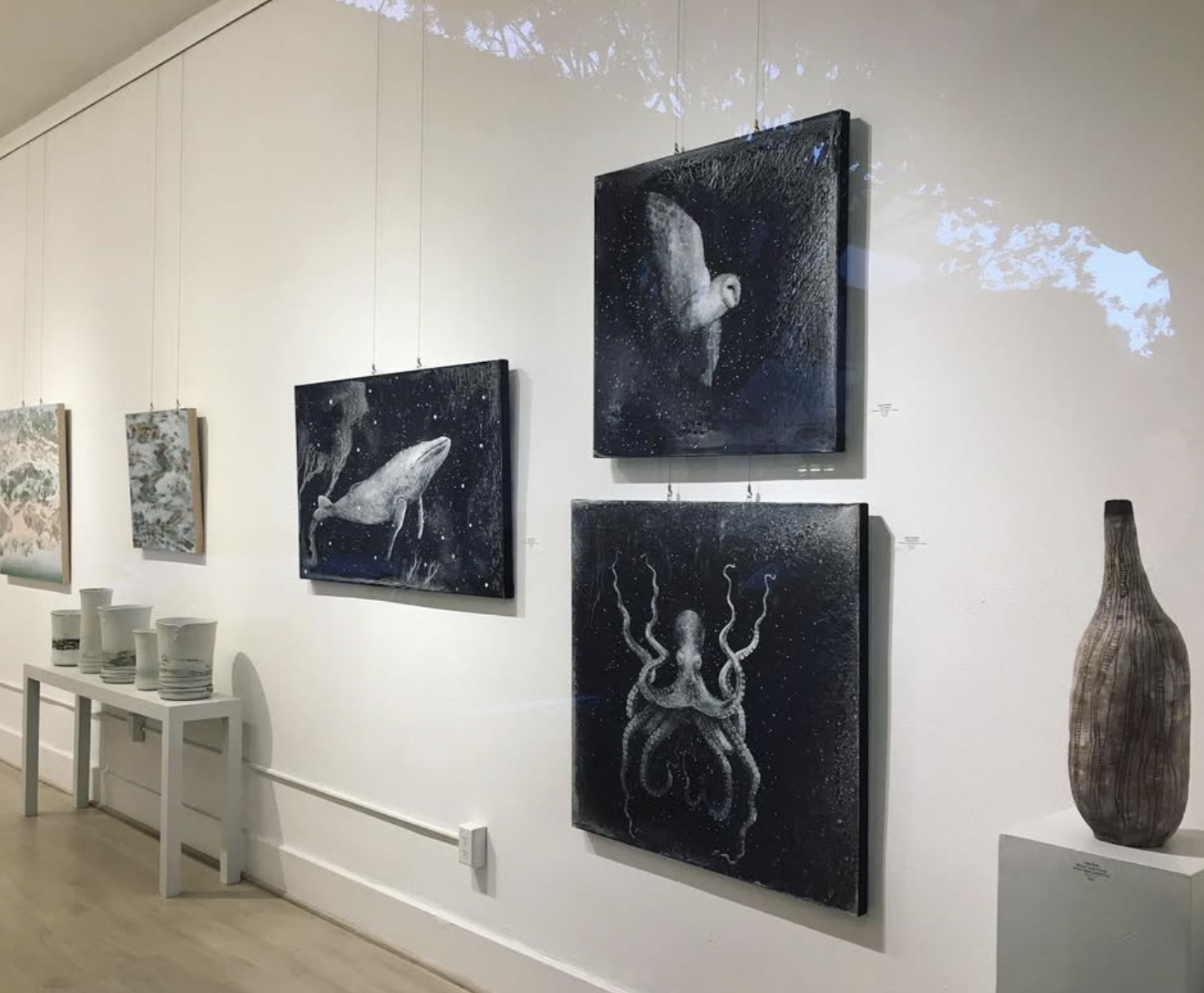 Art gallery wall with black and white ocean-themed paintings, a tall vase on a white pedestal, and white pottery on a white table.