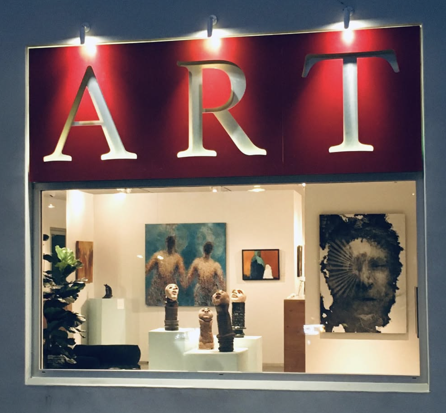 Art gallery window displaying sculptures on pedestals, paintings on the walls, and a red sign with large illuminated letters spelling 'ART'.