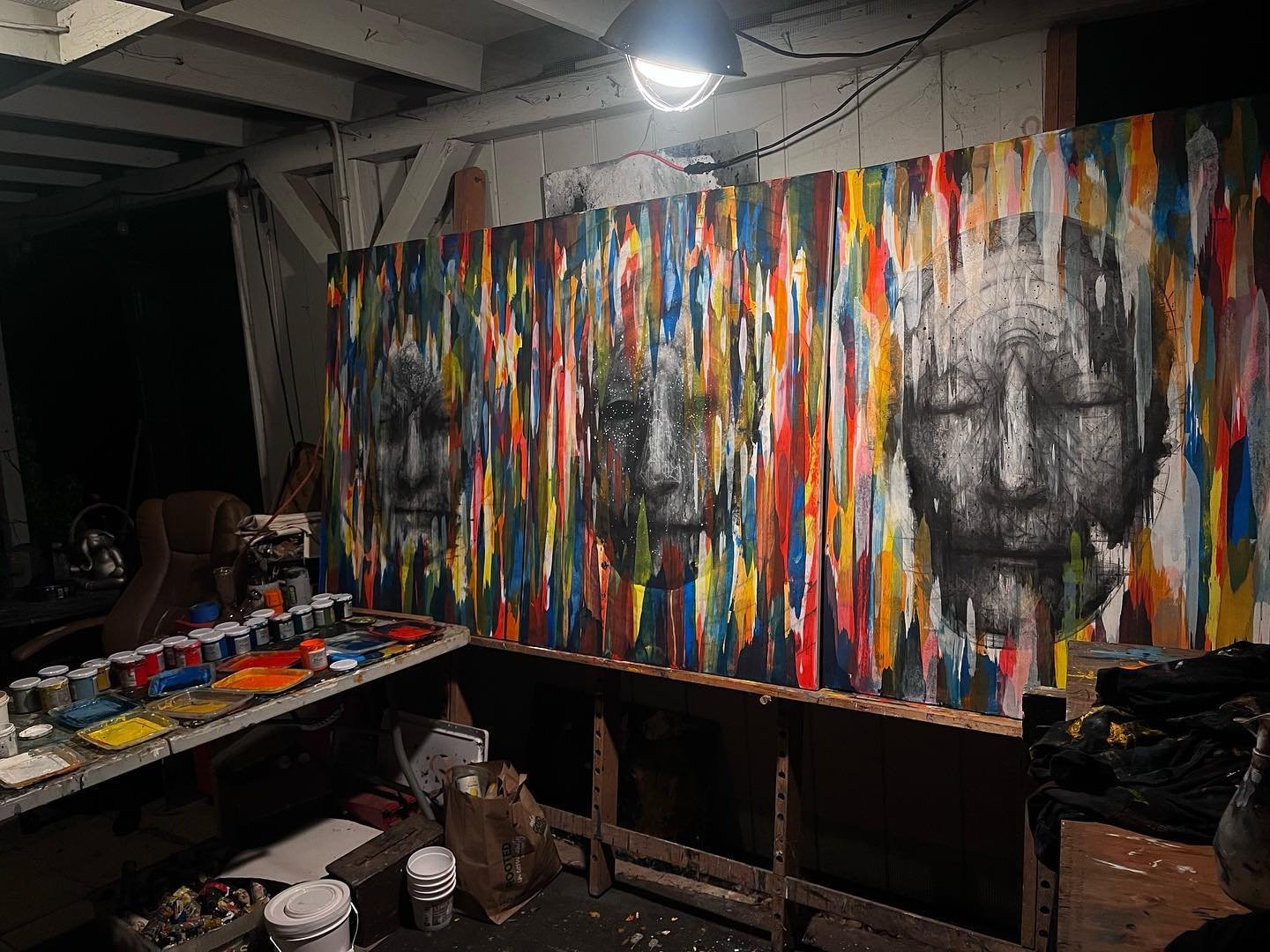 Three large abstract portraits painted on canvases with colorful drips and splashes, displayed on a wall in an art studio. A table in the foreground holds multiple small paint containers, brushes, and art supplies.