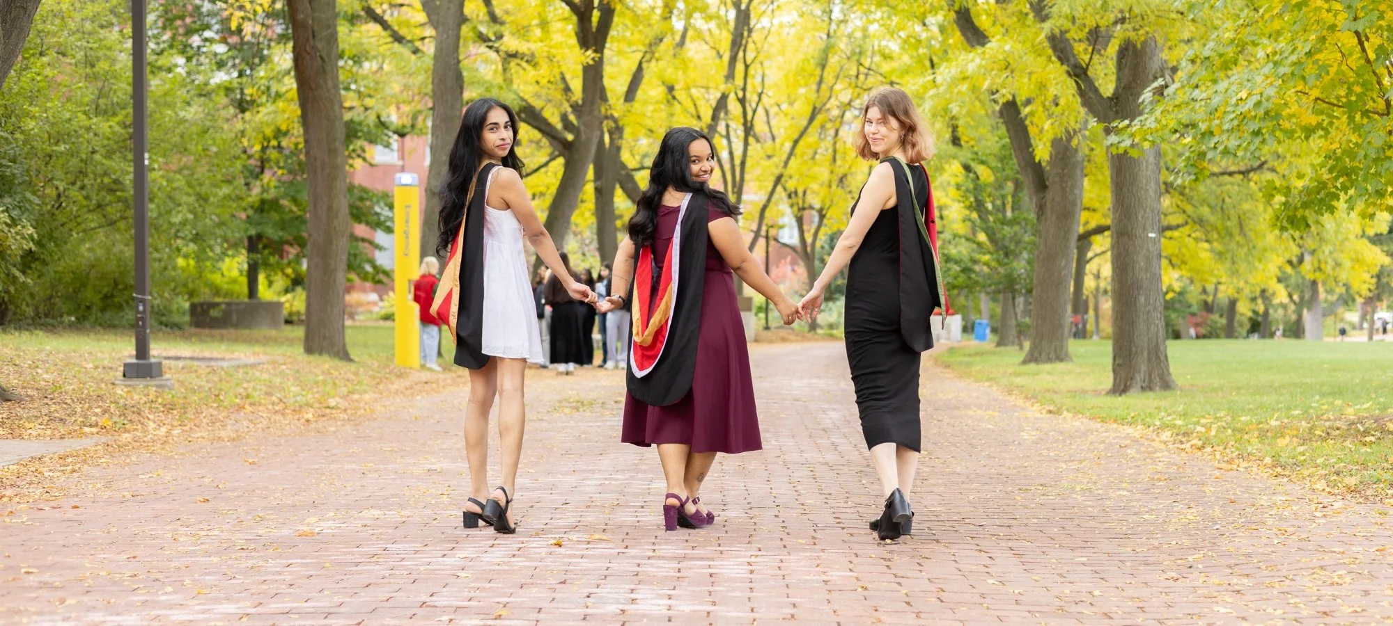 Three young women in graduation gowns holding hands and walking on a brick path in a park during fall.