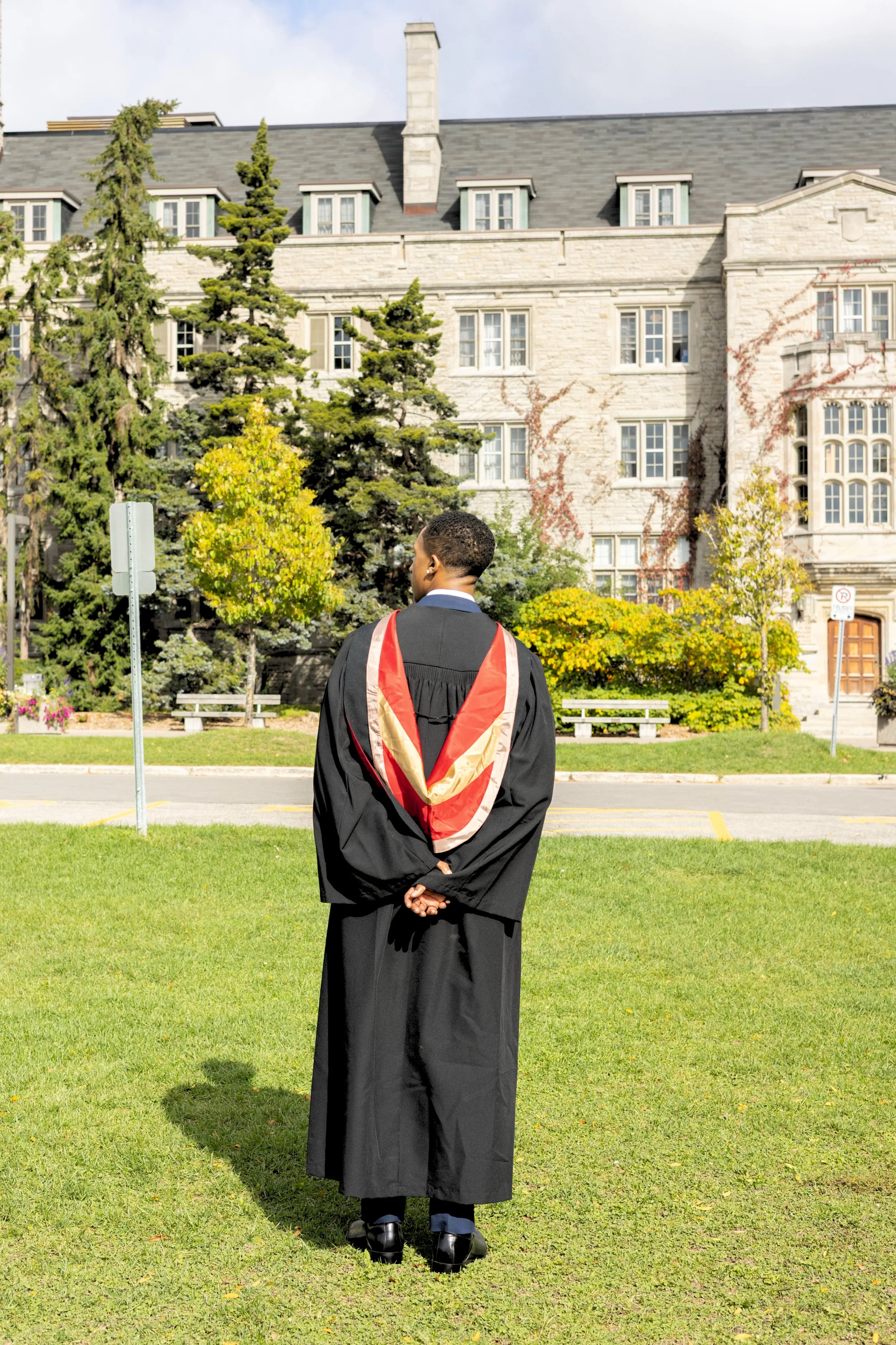 A man dressed in graduation gown standing on a grass lawn in front of a large historic building with trees and bushes.