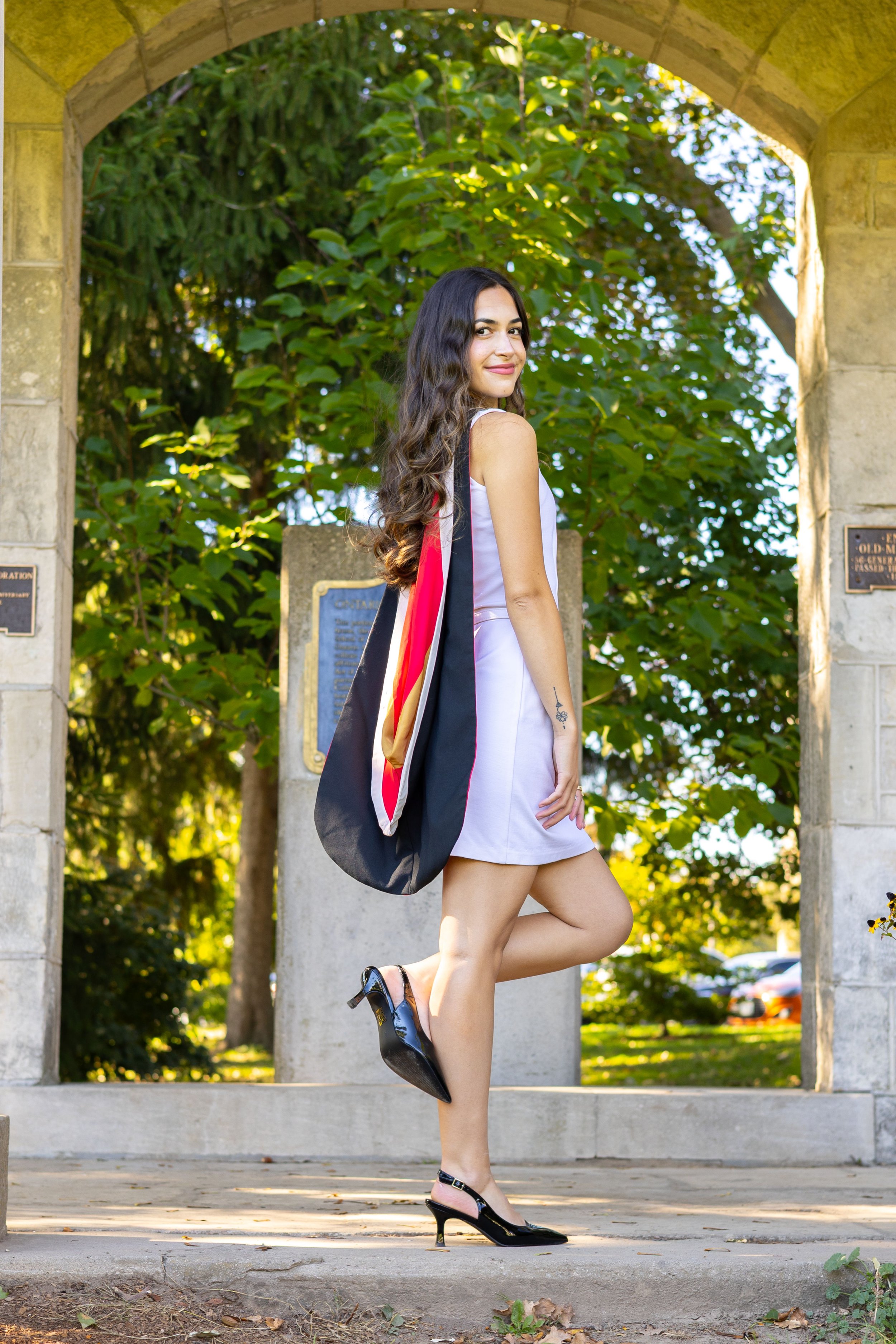 A young woman in a white dress and black high heels stands happily under an archway, holding a black tote bag with red and gold accents. She has long, wavy brown hair and is standing on one leg with the other slightly bent, smiling at the camera. Lus