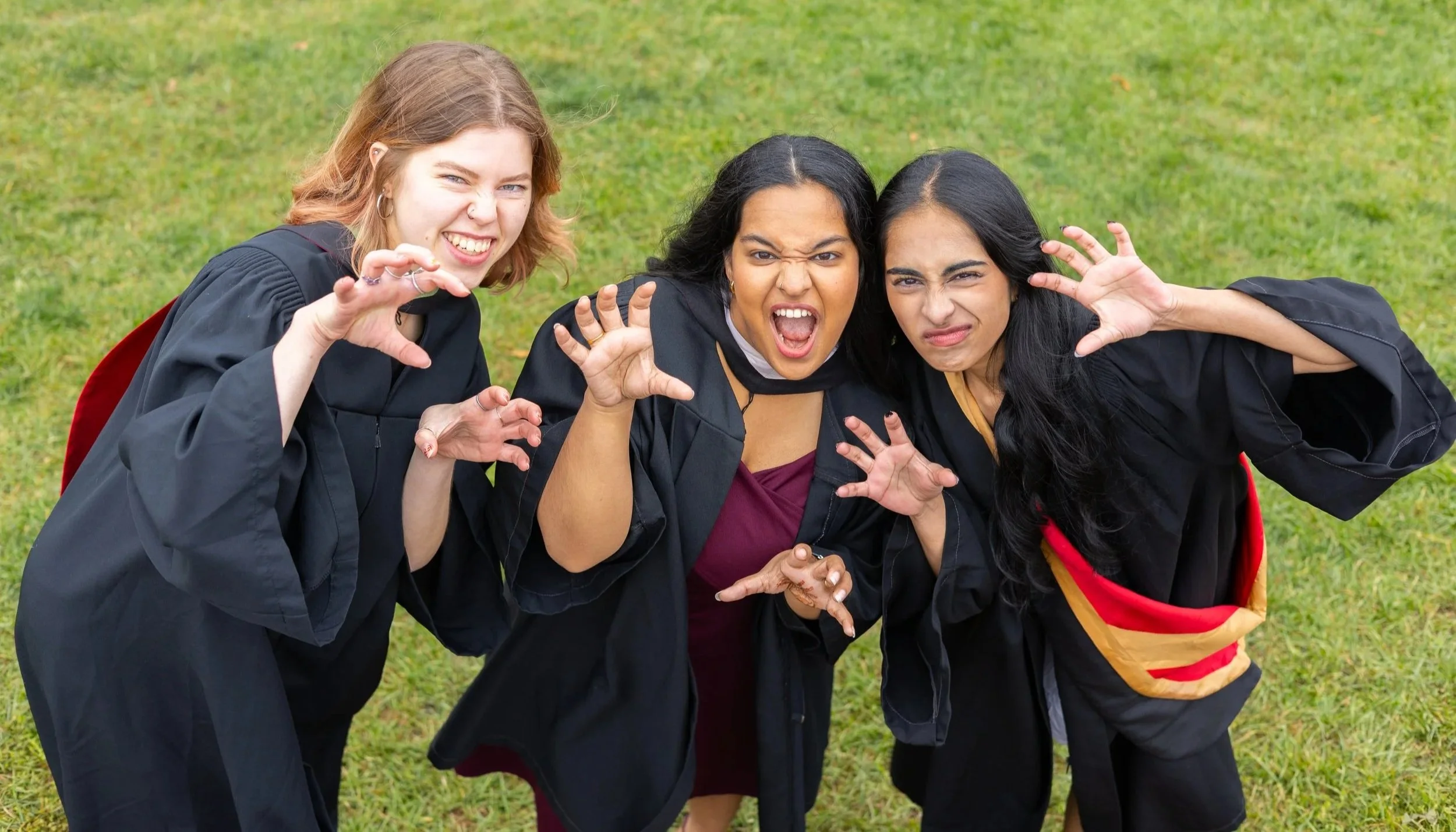 Three young women in graduation gowns making funny faces and pretending claws while crouching on grass.
