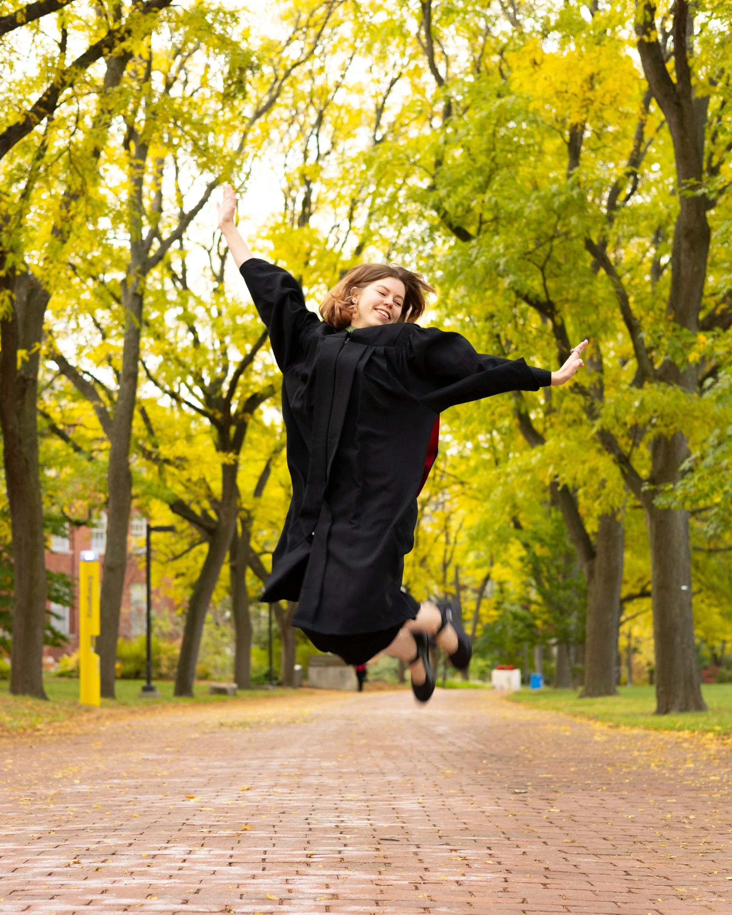 A young woman in a graduation gown joyfully jumping in a park with yellow and green autumn leaves.