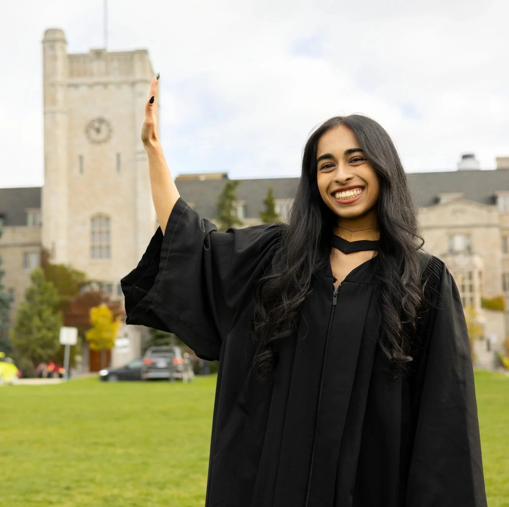 A smiling woman in a graduation gown standing outdoors in front of a historic building with a tower and clock, holding up her hand as if waving or about to toss a cap.