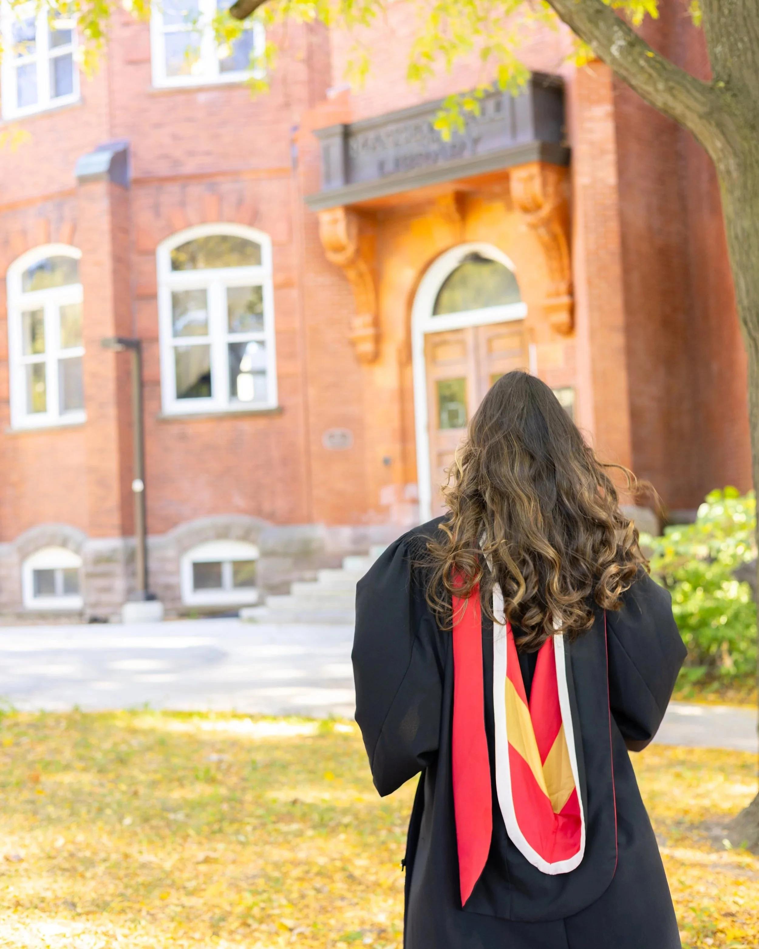 Person with long curly hair and a graduation gown standing outside in front of red brick building with arched windows and steps.