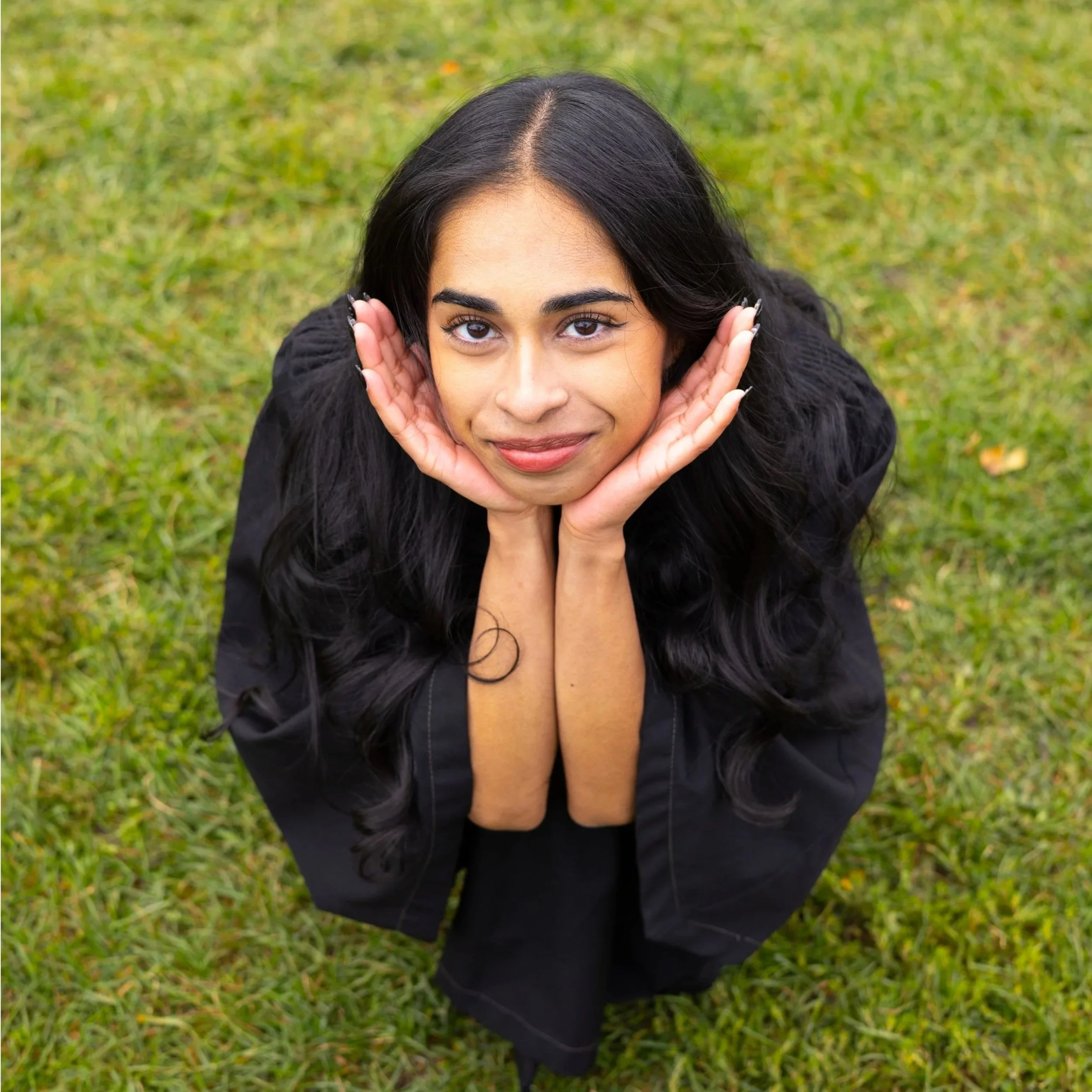 A young woman with long black hair and a black jacket looks up at the camera while resting her face in her hands, outdoors on a grassy field.