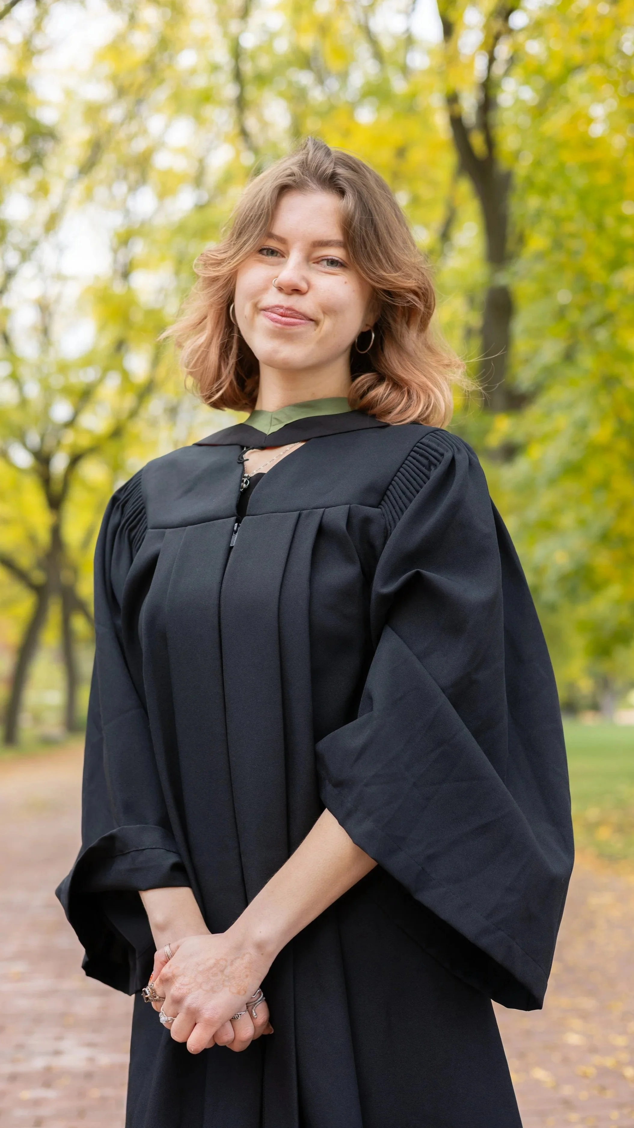 A young woman standing outdoors in a graduation cap and gown, smiling with trees and autumn leaves in the background.
