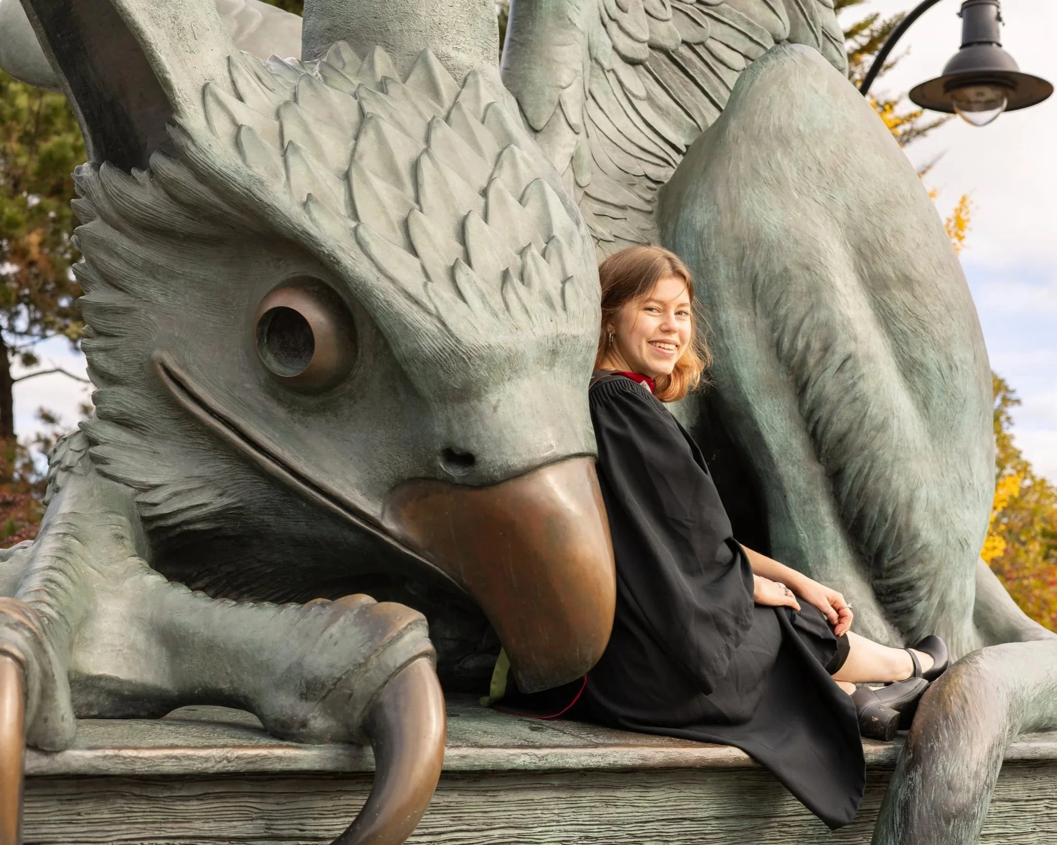 A young woman with blonde hair, smiling, sitting on a large bronze sculpture of a dragon with a bird's head. The sculpture features detailed scales and large wings. The background has trees with autumn foliage and a lamp.