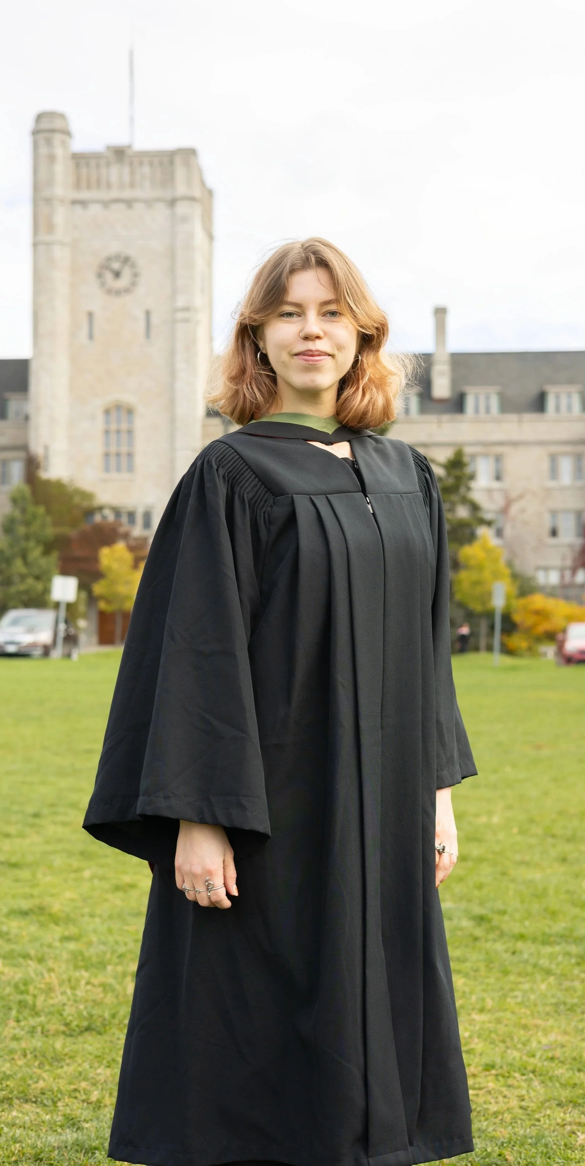 Young woman in a black graduation gown standing on a green lawn with a historic building in the background.