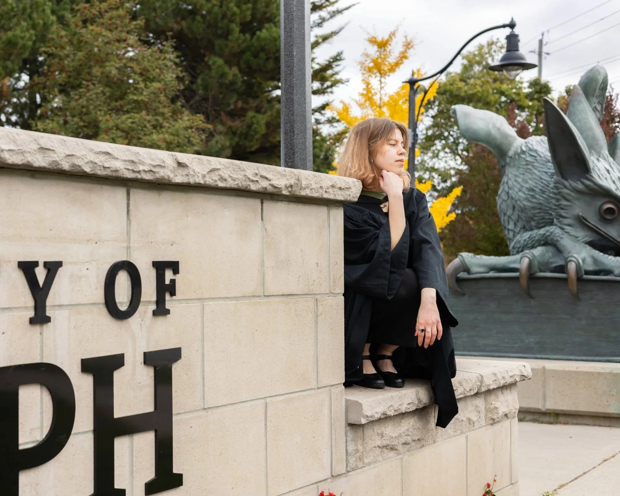 A young woman dressed in a black gown sitting on a stone ledge next to a sculpture of a fish with a large open mouth, behind a stone wall with partially visible text reading 'OY OF PHIL', in an outdoor park with trees displaying fall foliage.
