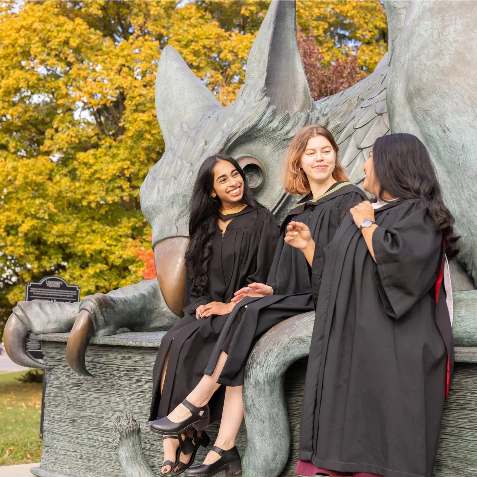 Three women in graduation gowns sitting on a bench shaped like a dinosaur, with autumn trees in the background.