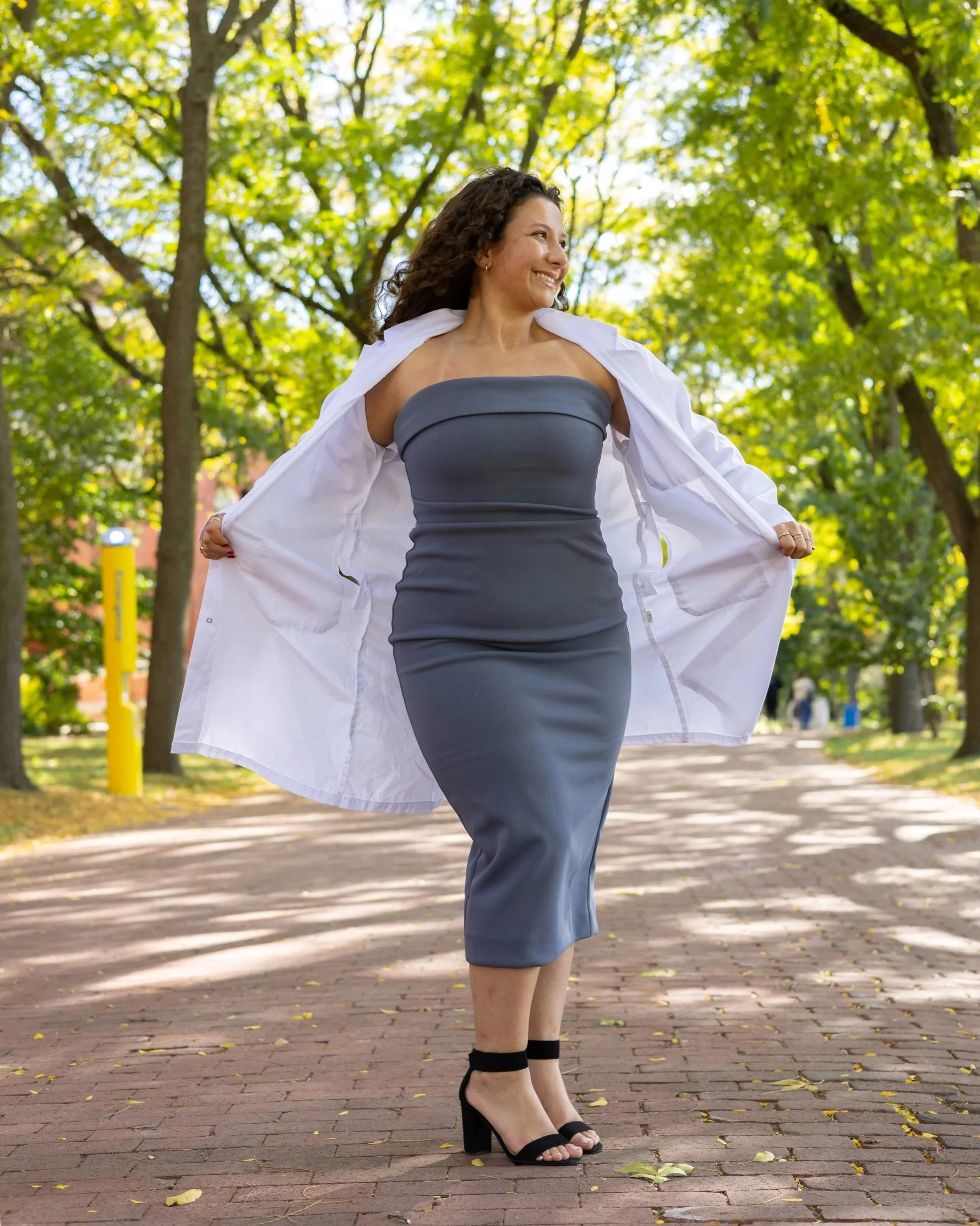 A woman in a gray strapless dress and black heels walking outdoors with a white coat draped over her shoulders, smiling and holding the coat open in a park with green trees and a brick pathway.