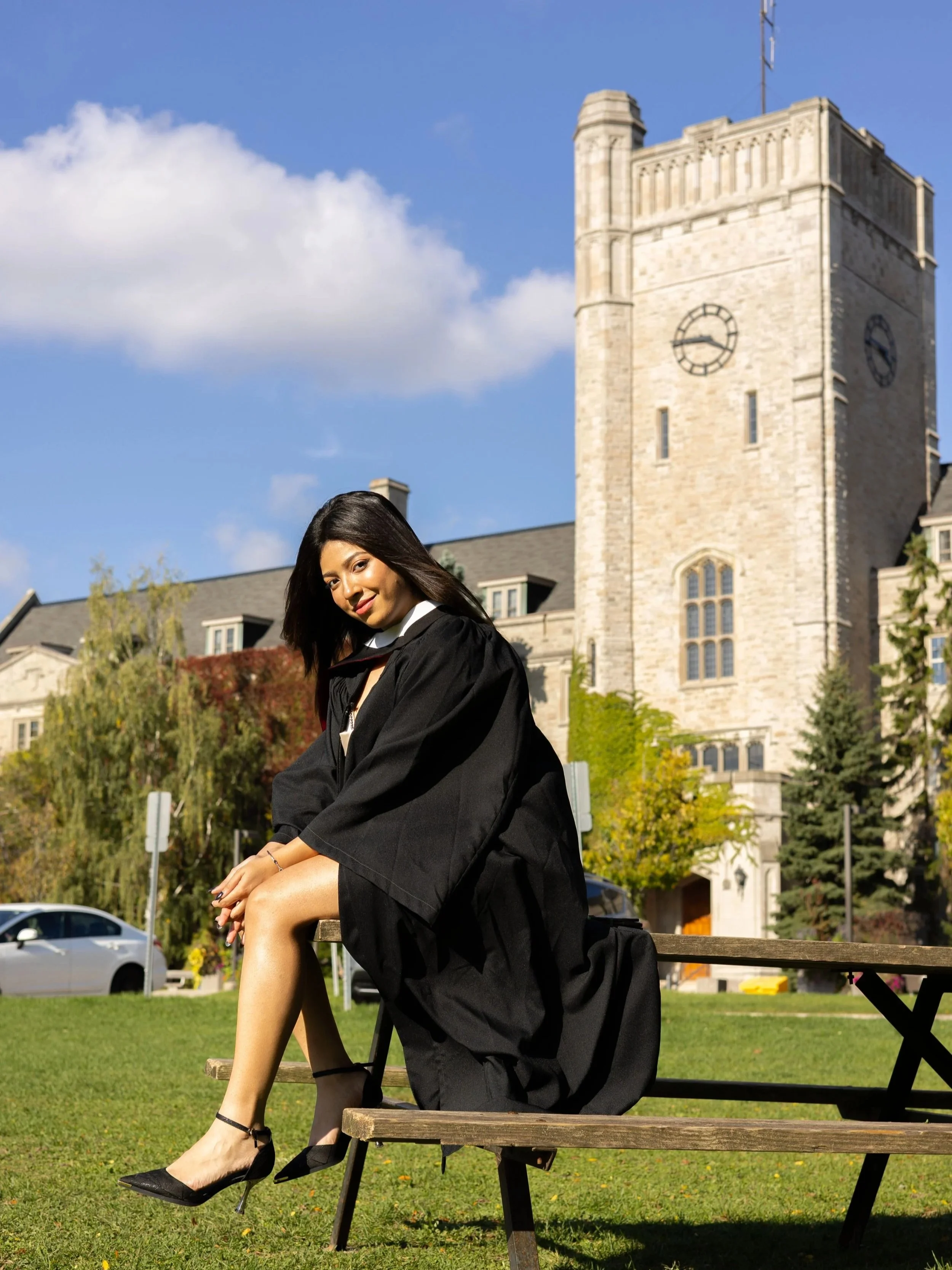 A woman in graduation gown sitting on a park bench with a historic building in the background, a stone clock tower, and a clear blue sky.